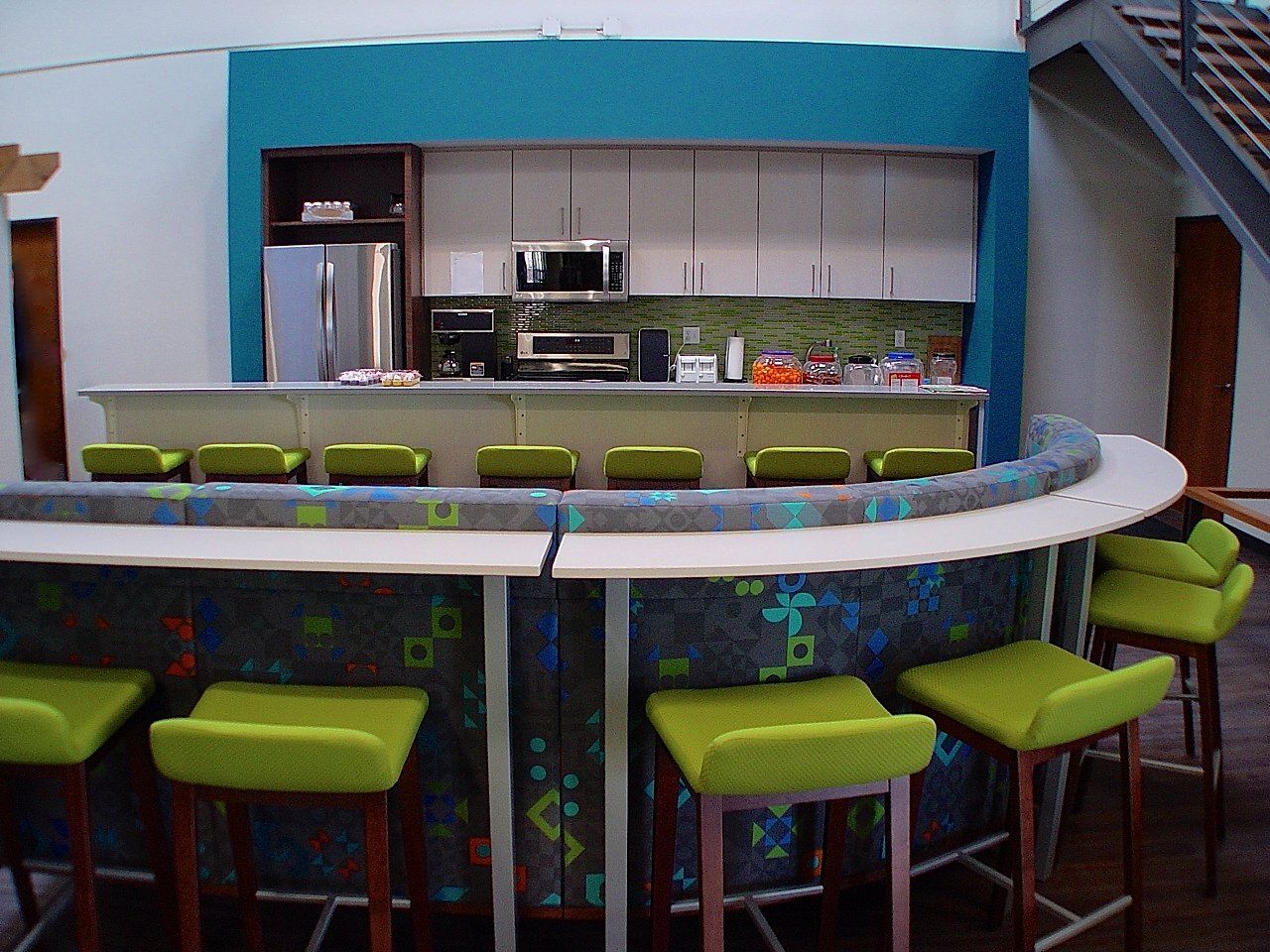 Bar with curved counter and green stools in front of a turquoise wall with kitchen appliances.