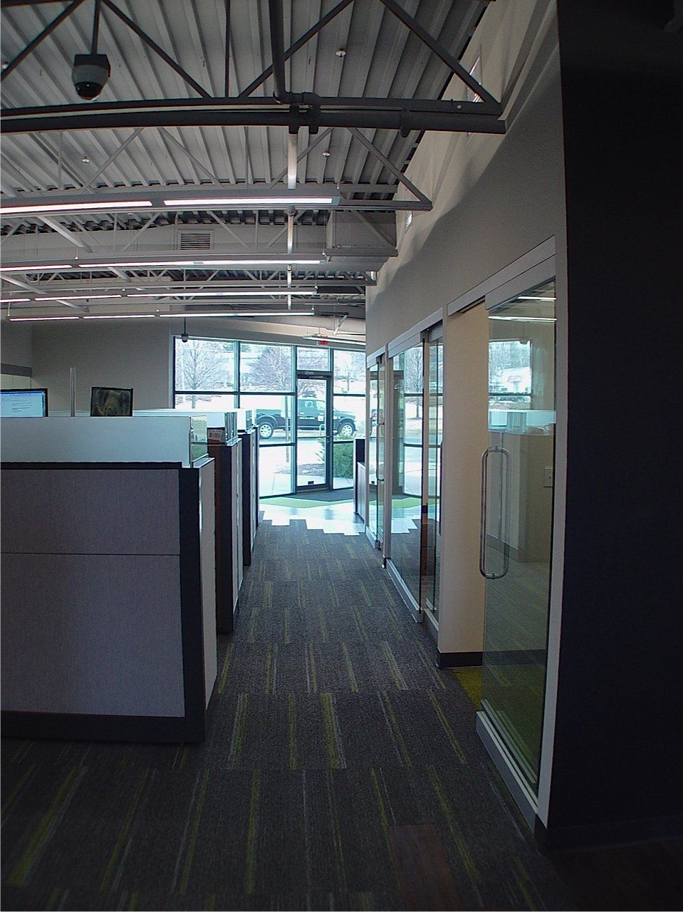 Office hallway with cubicles on the left and glass-walled offices on the right. Natural light streams in from the end.