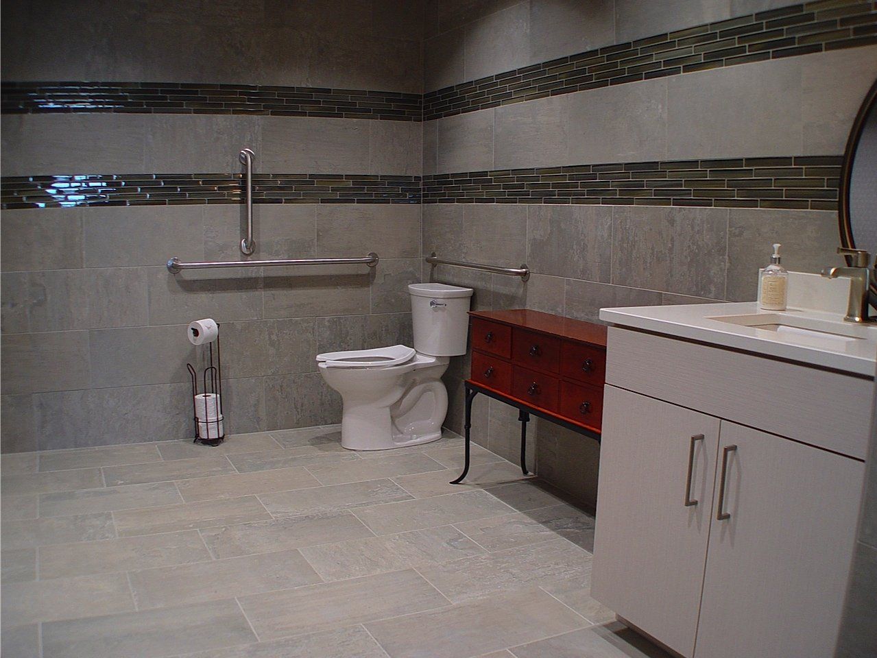 Accessible bathroom with grab bars, toilet, and sink. Neutral gray tiles and brown cabinets.