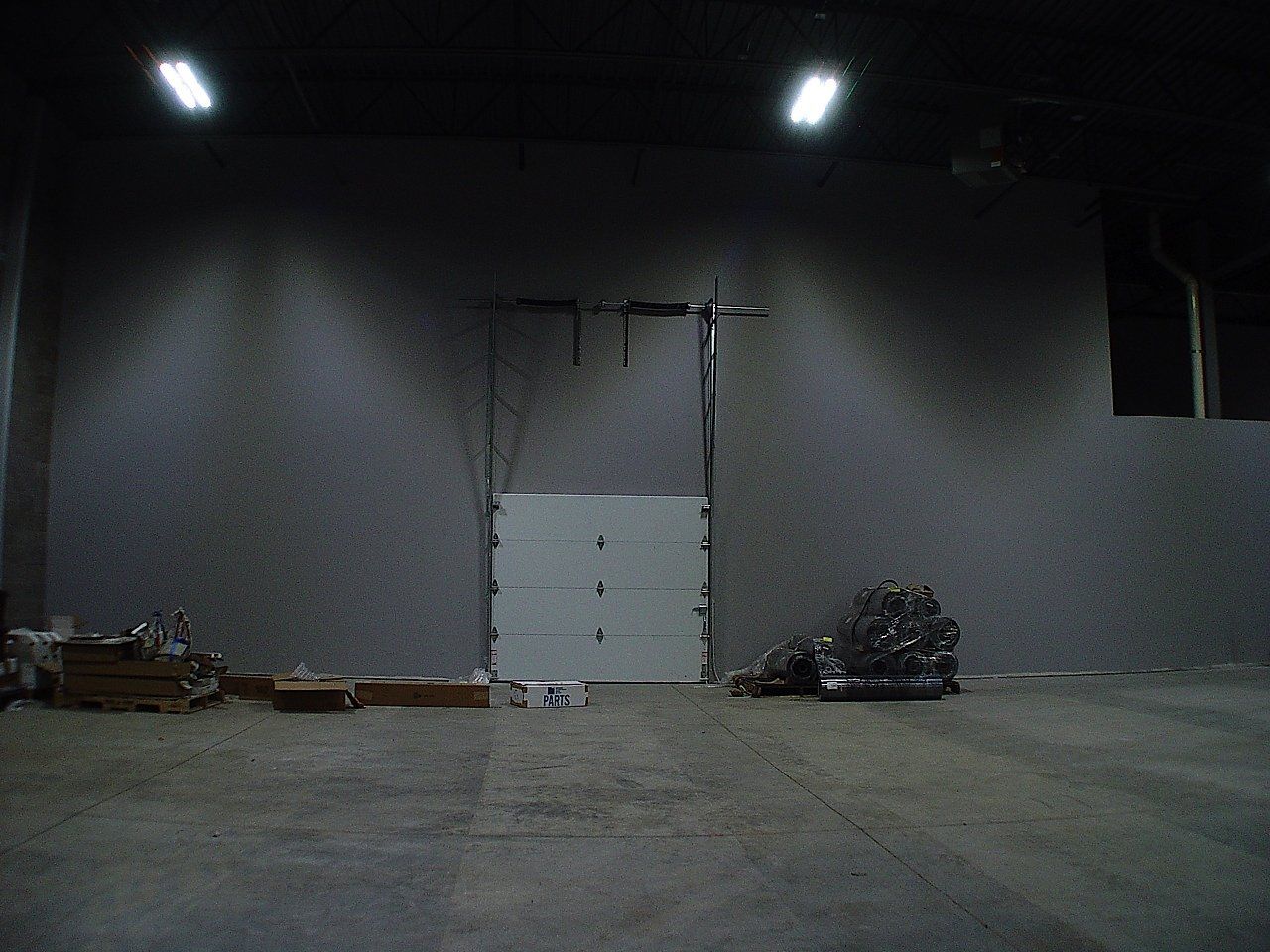 Interior view of a gray industrial space with a closed garage door. Fluorescent lights illuminate the upper area; objects lie on the floor.