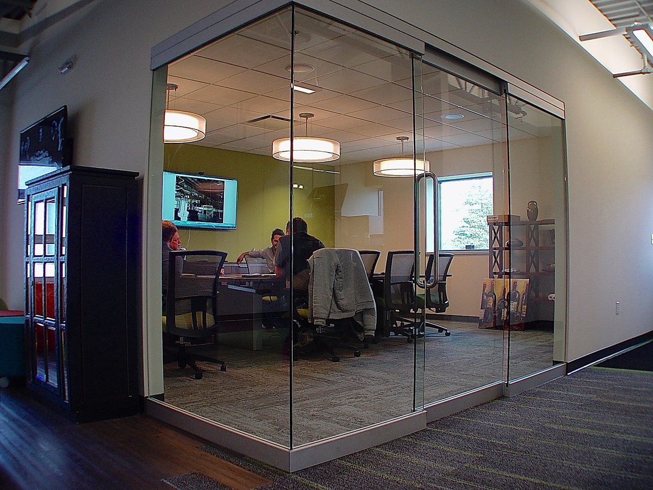 Conference room with glass walls, people at a table, and a TV screen.