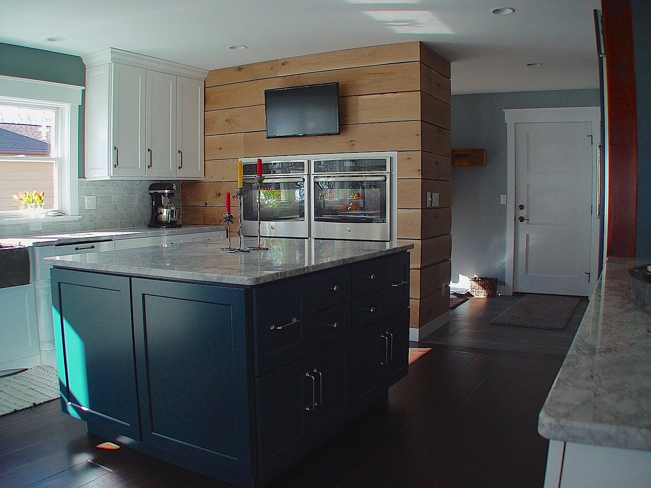 Modern kitchen with navy blue island, white cabinets, and wood-paneled wall with ovens and small TV.