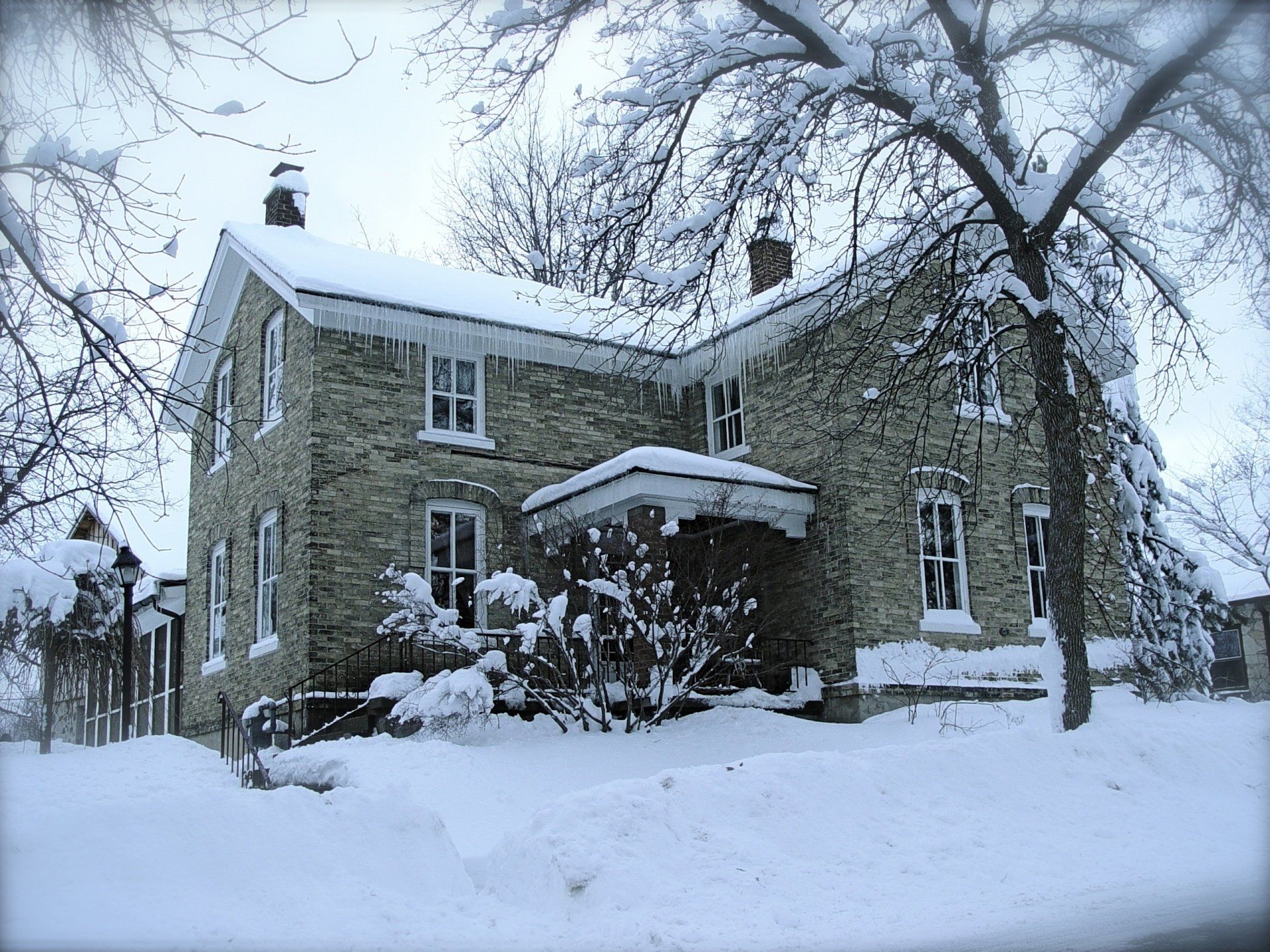 Snow-covered stone house with icicles hanging from the roof. Winter scene with snowy ground and bare trees.