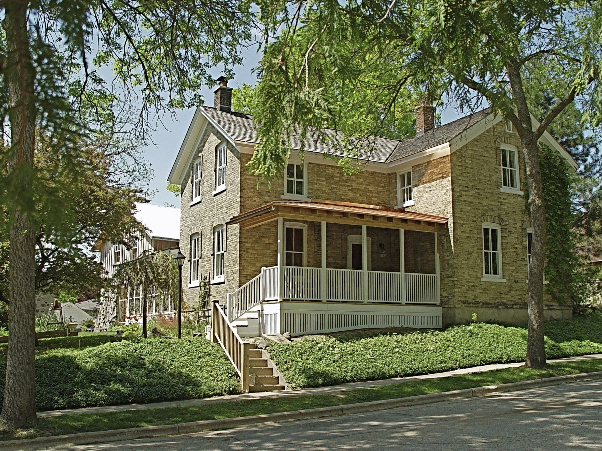 Yellow brick two-story house with porch and stairs, green lawn, and trees on a sunny day.