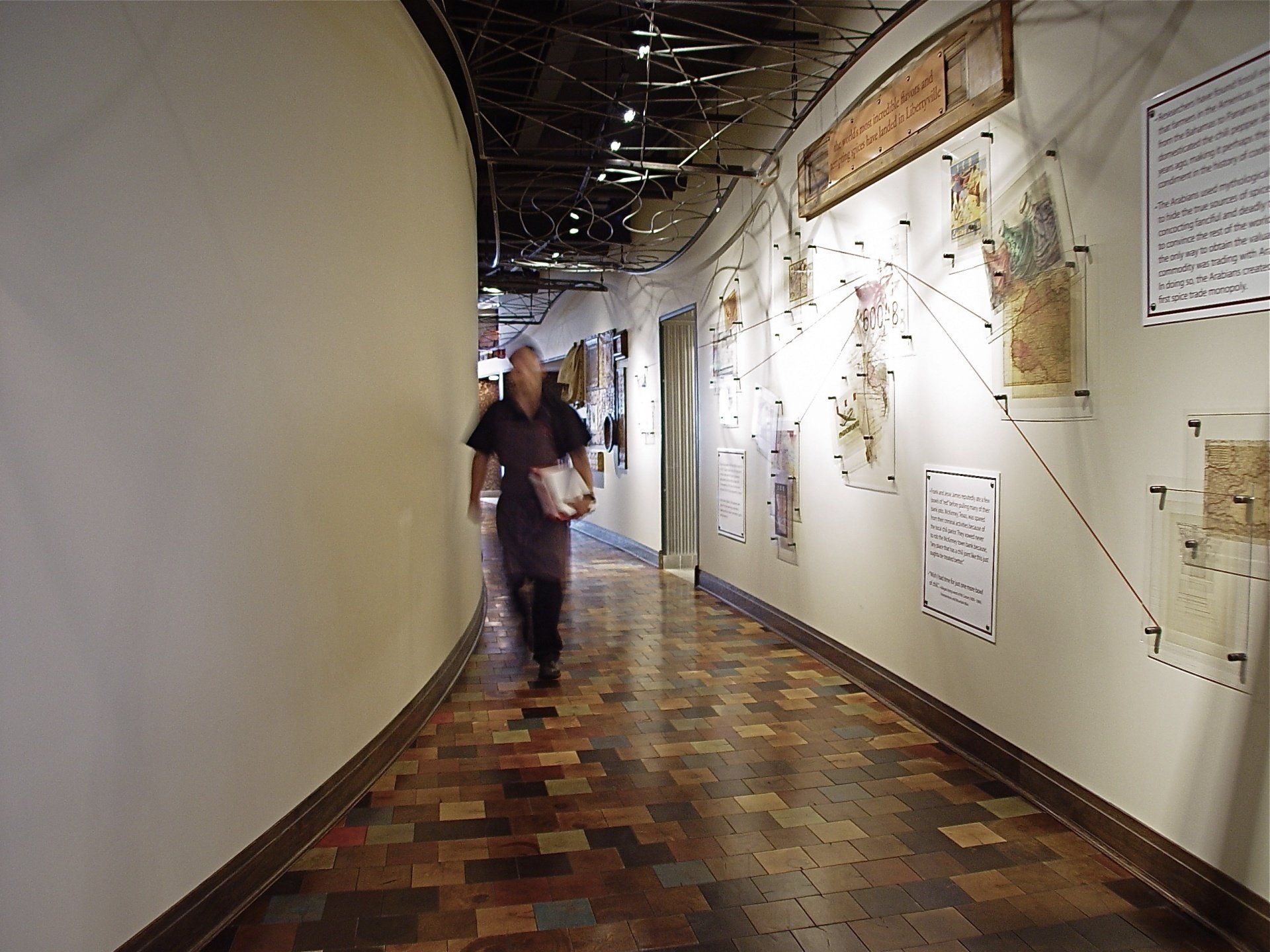 Person walking in a museum hallway with displays on the right wall. The floor has a brick pattern.
