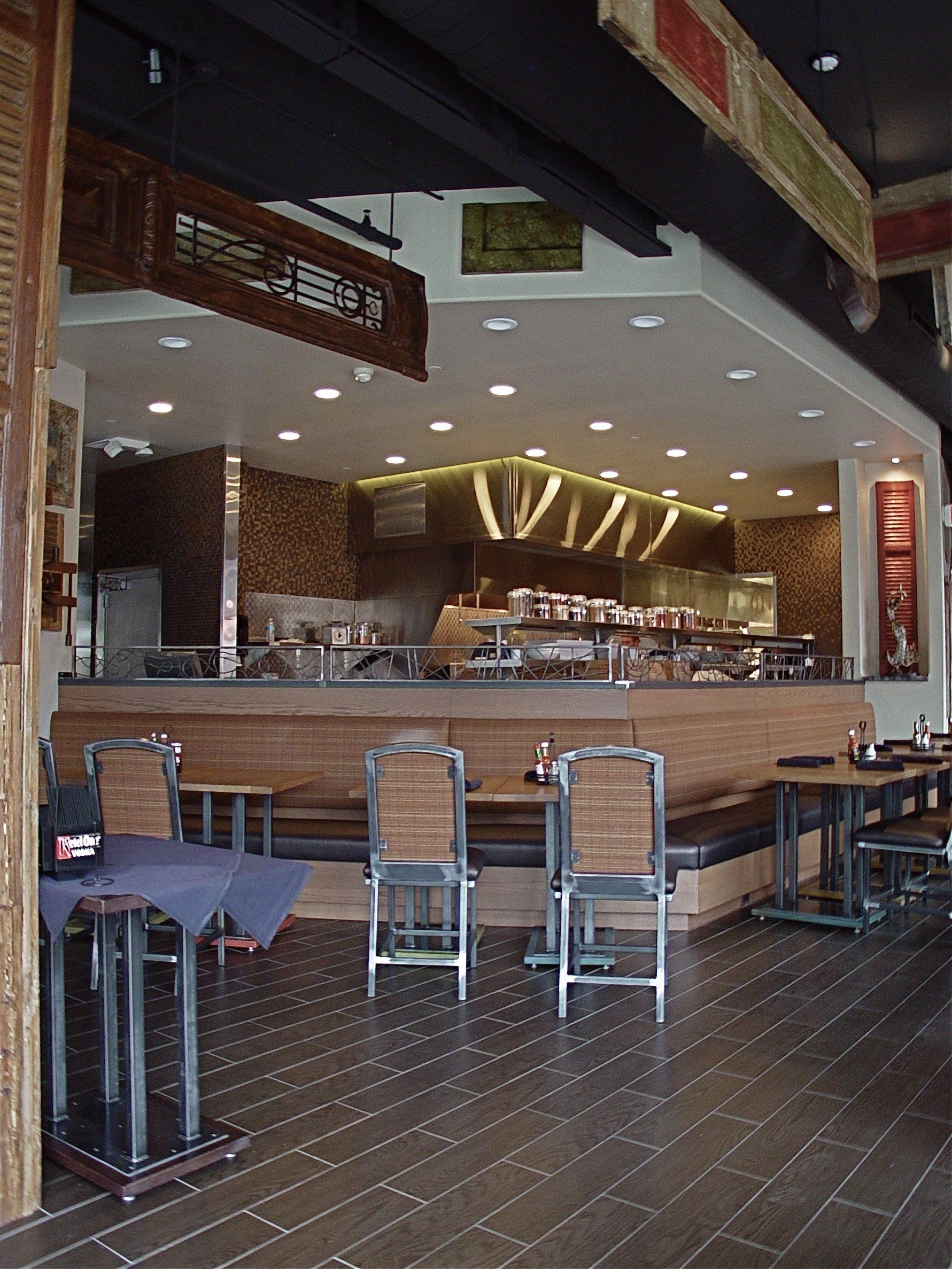 Restaurant interior with wooden floor and seating, view of the counter and kitchen, overhead lighting.