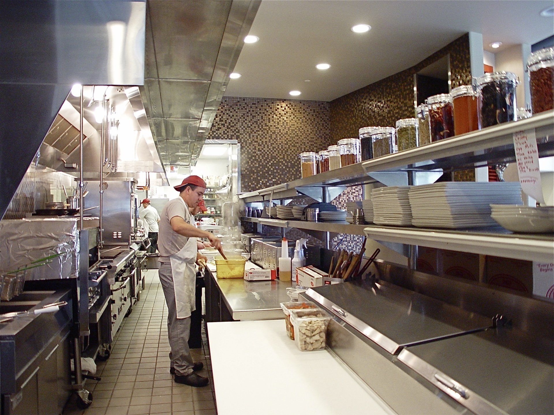 Kitchen worker cooking at a restaurant. Stainless steel appliances, shelves of spices, and plates.