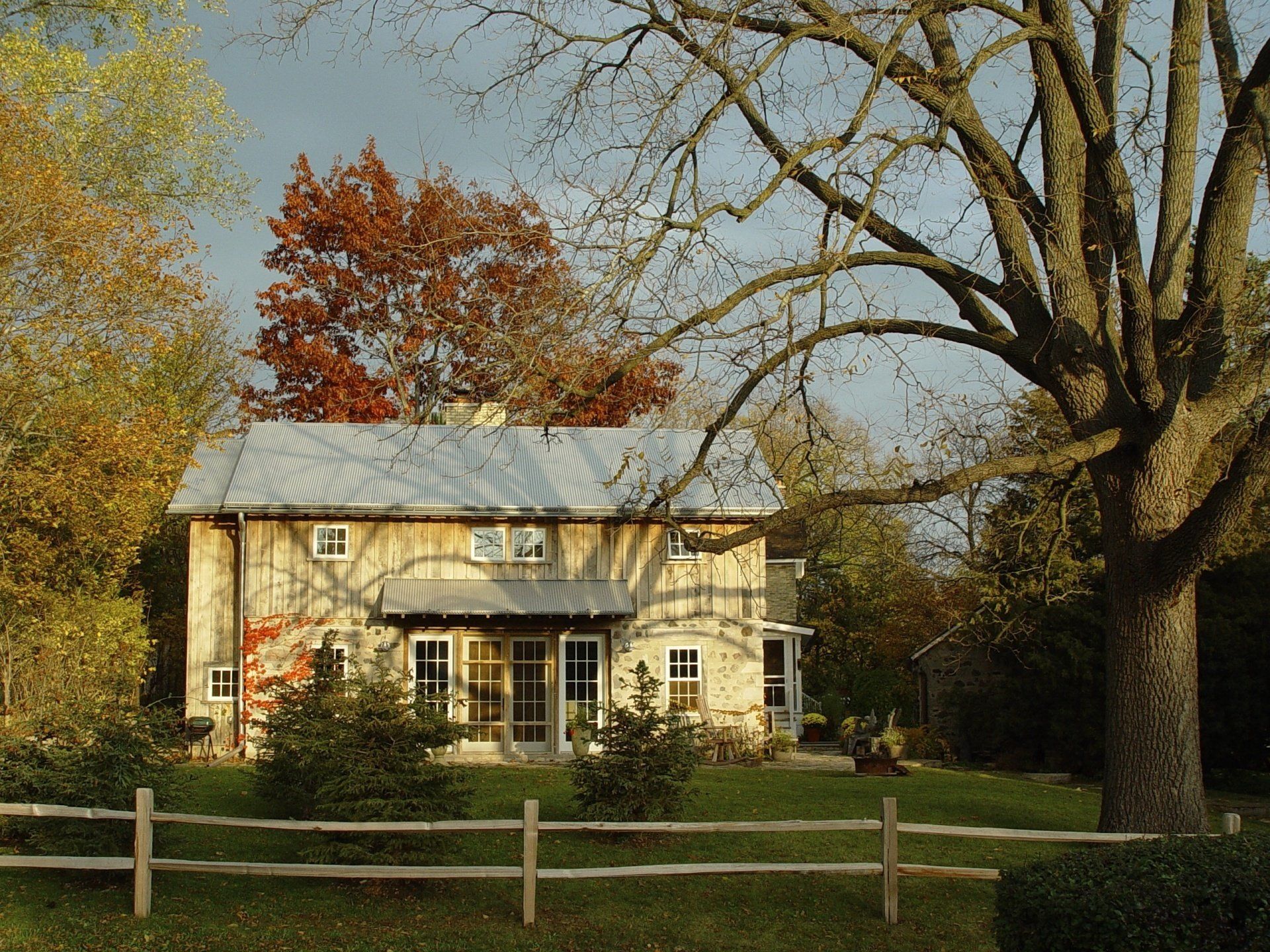 Two-story wooden house with a metal roof and porch, set amongst fall foliage. A split-rail fence in the foreground.