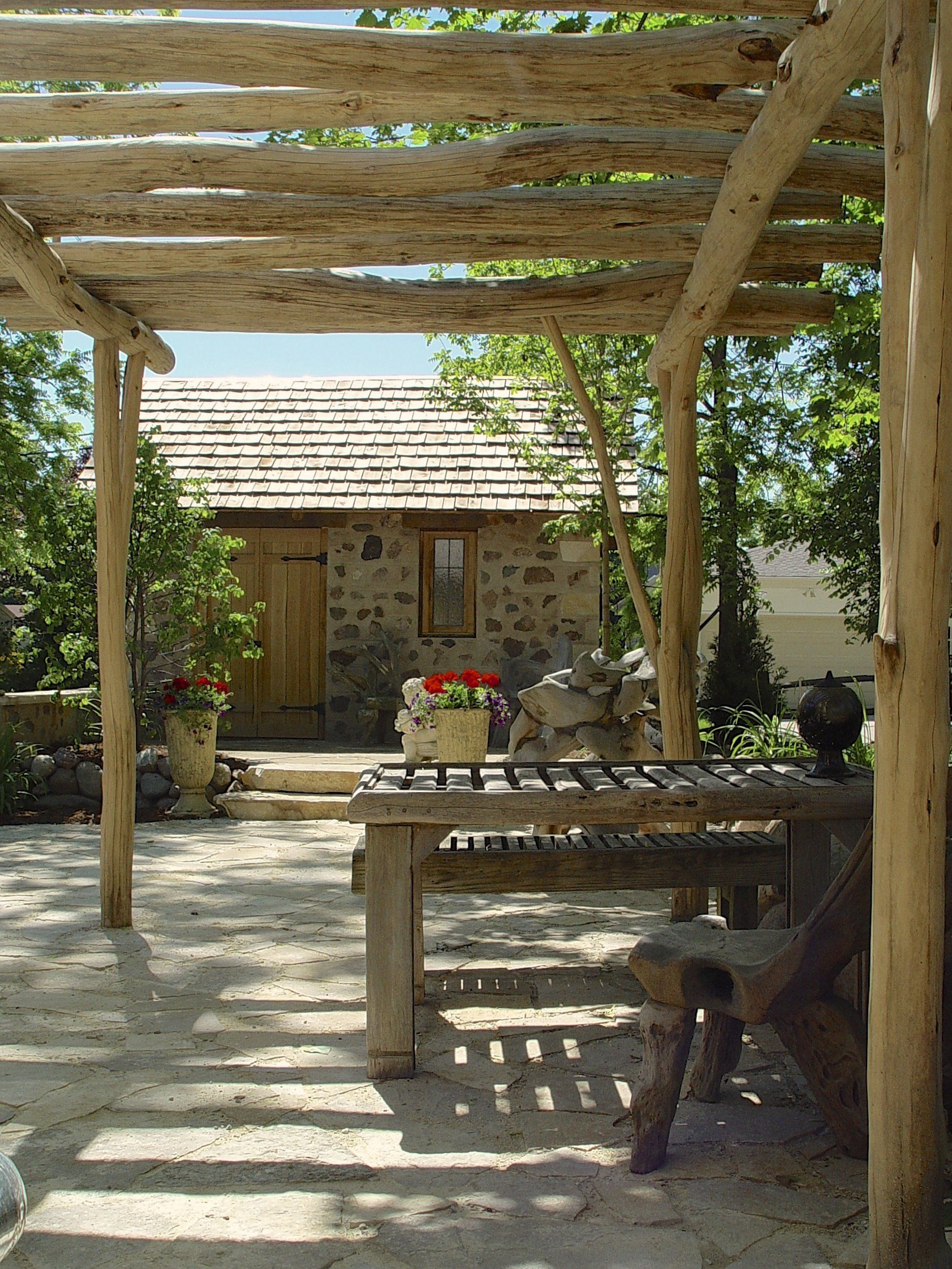 Rustic wooden pergola over a stone patio with a table and a small stone building in the background.