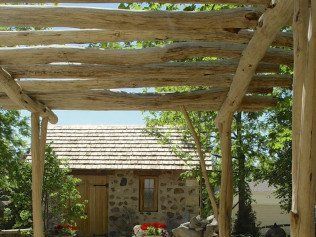 Wooden pergola with a view of a stone building with a wood-shingled roof.