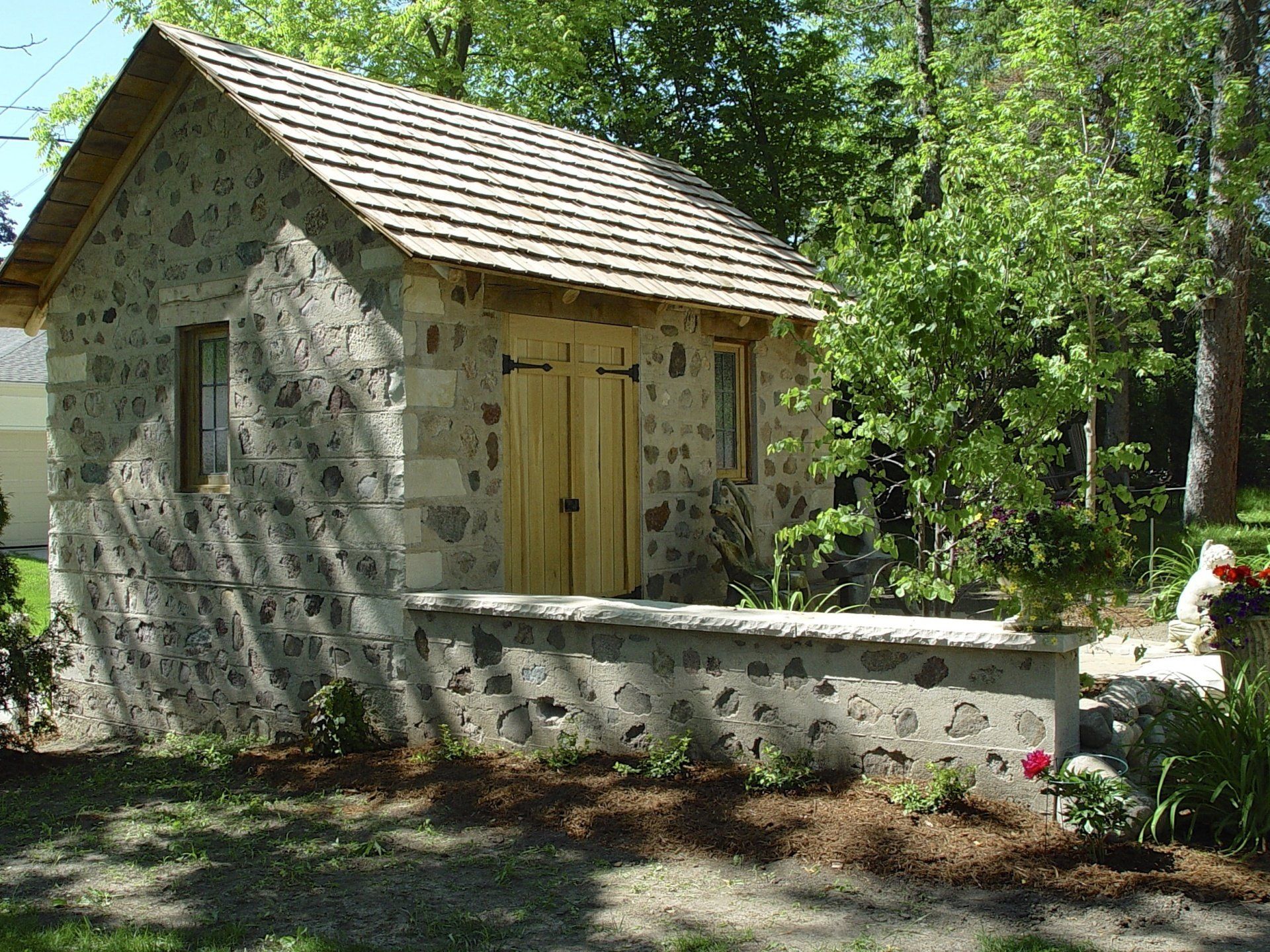Stone shed with wooden door and roof, small windows, surrounded by trees and a stone wall.