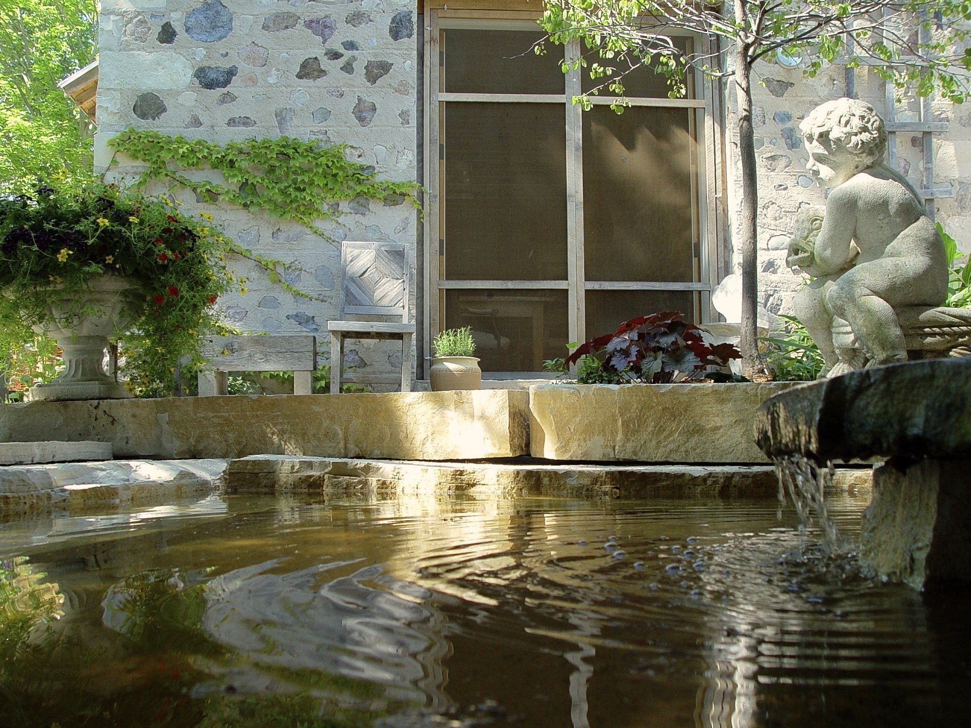 Stone building with a reflecting pool and a cherub statue. Green plants and flowers decorate the area.