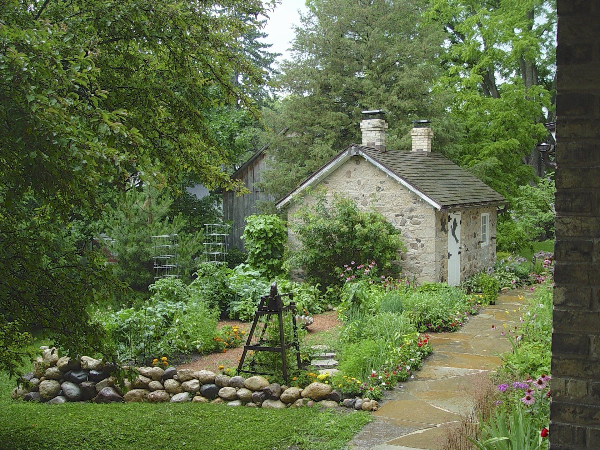 Stone cottage in a lush garden with a pathway and vegetable beds. Trees and greenery surround the cottage.