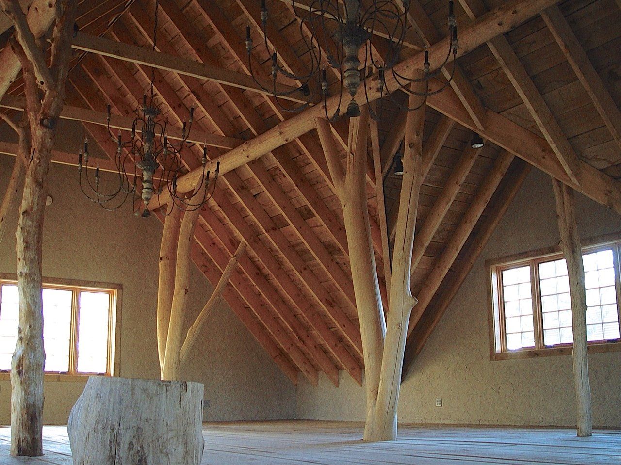 Interior of a wooden structure with exposed beams, rafters, and tree trunk supports. Two windows let in light.