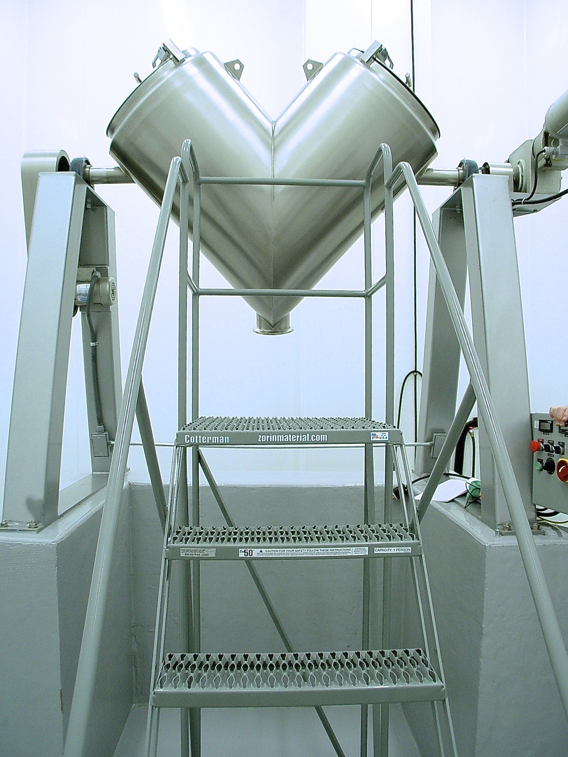 Stainless steel V-blender with access stairs, used in pharmaceutical or chemical processing.