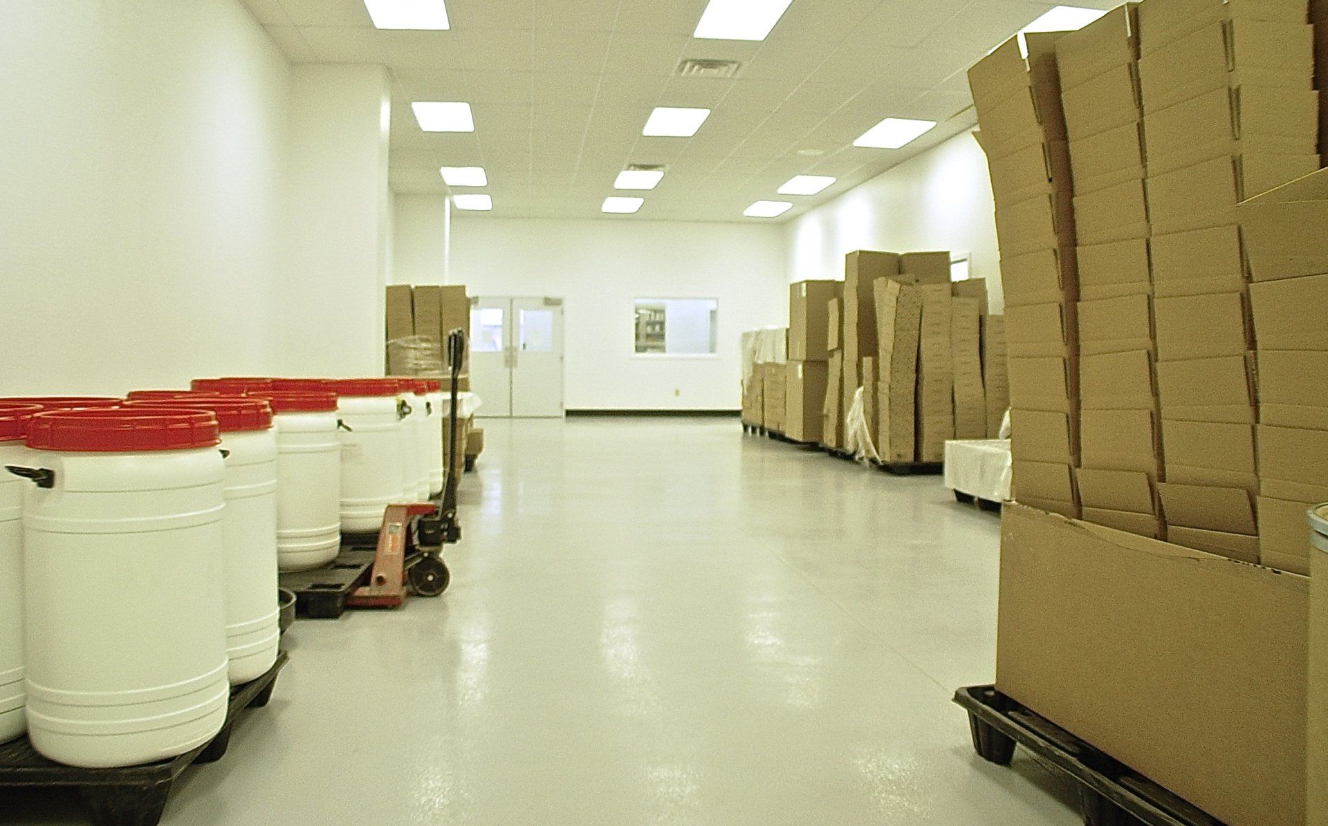 Warehouse interior with white walls, ceiling lights, and stacks of boxes and containers.