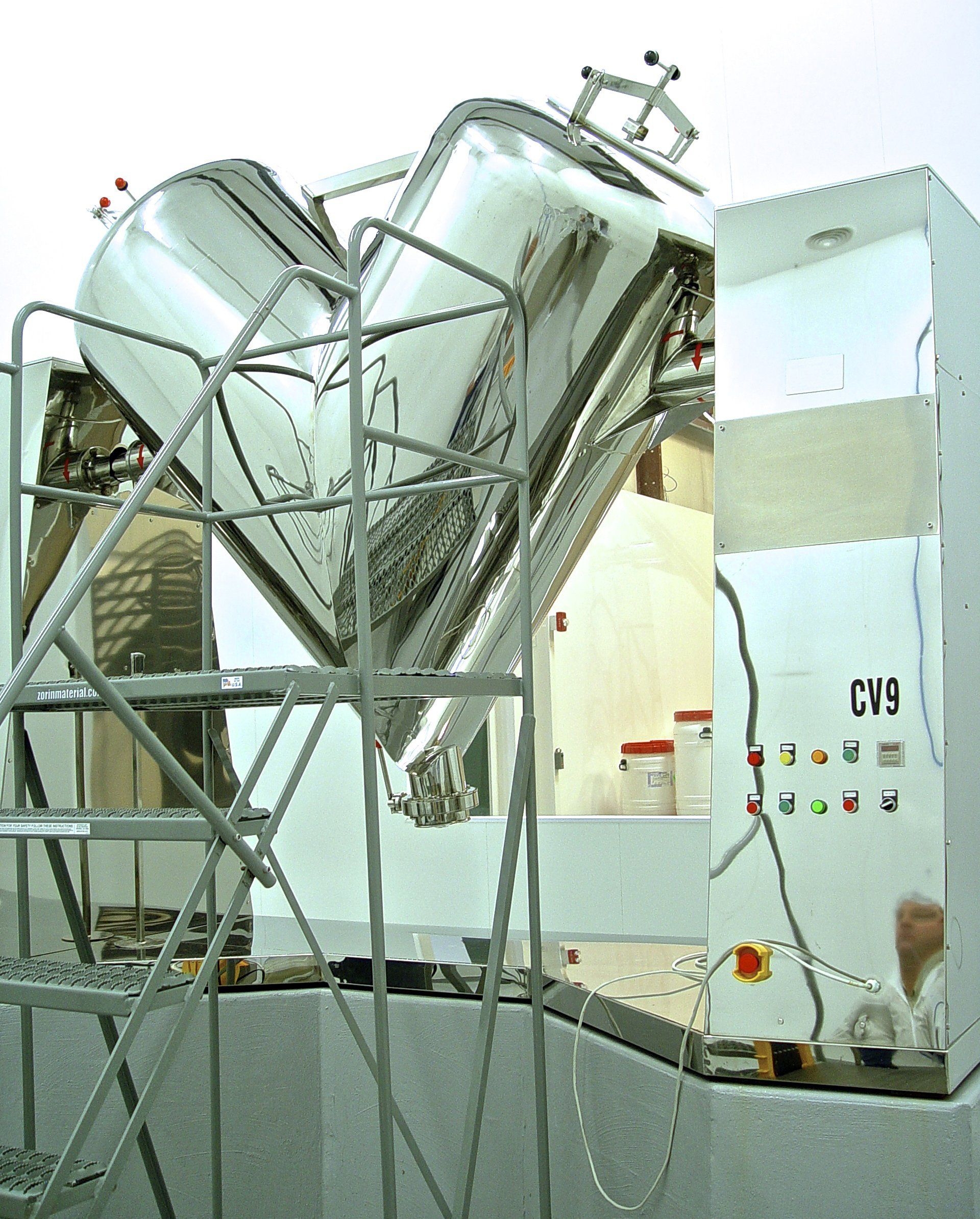 Stainless steel V-shaped blender inside a laboratory, mounted on a support structure.