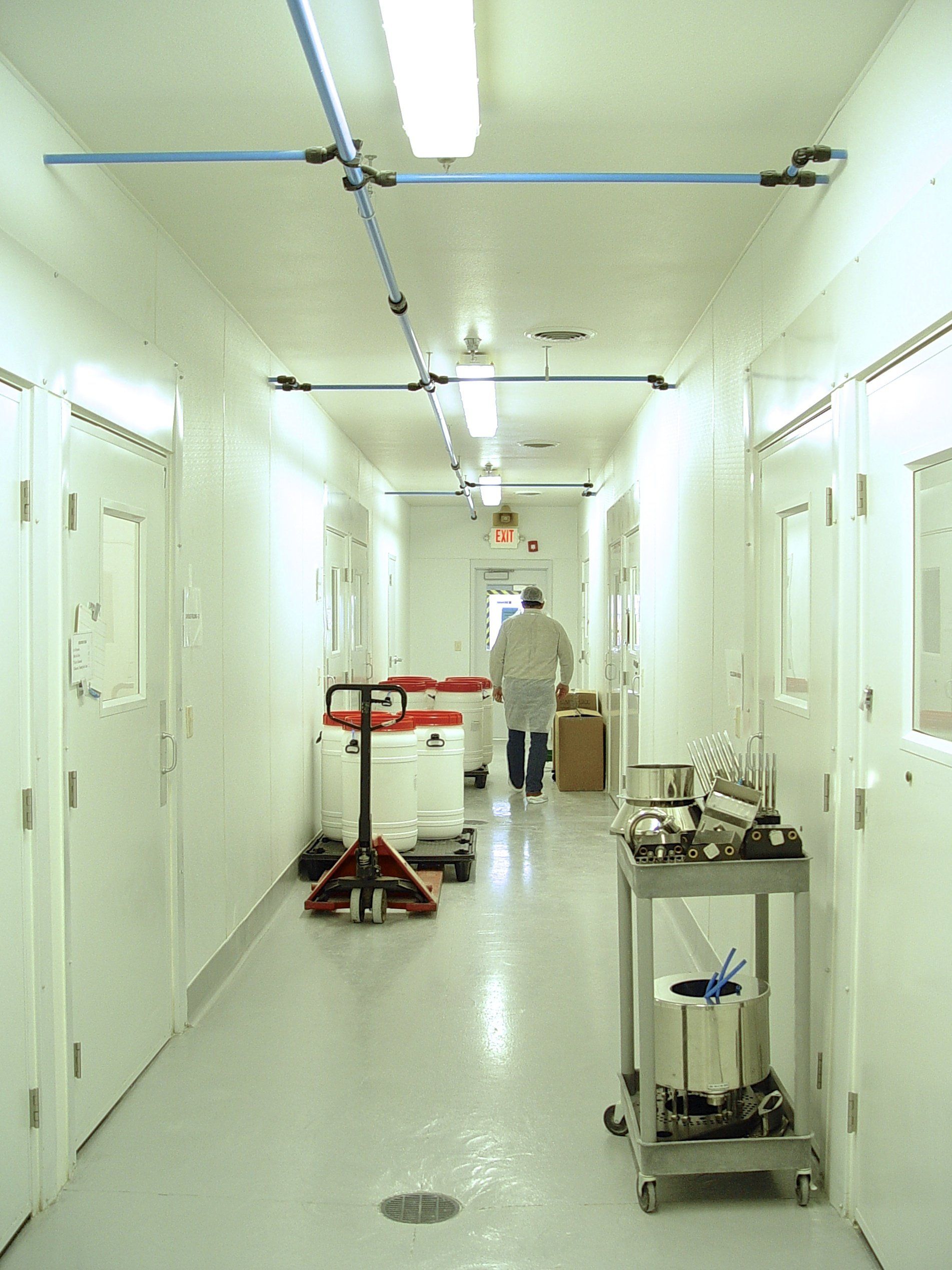 Laboratory hallway with white walls, doors, and floor; a person walks toward the end. Equipment on floor. Blue pipes overhead.