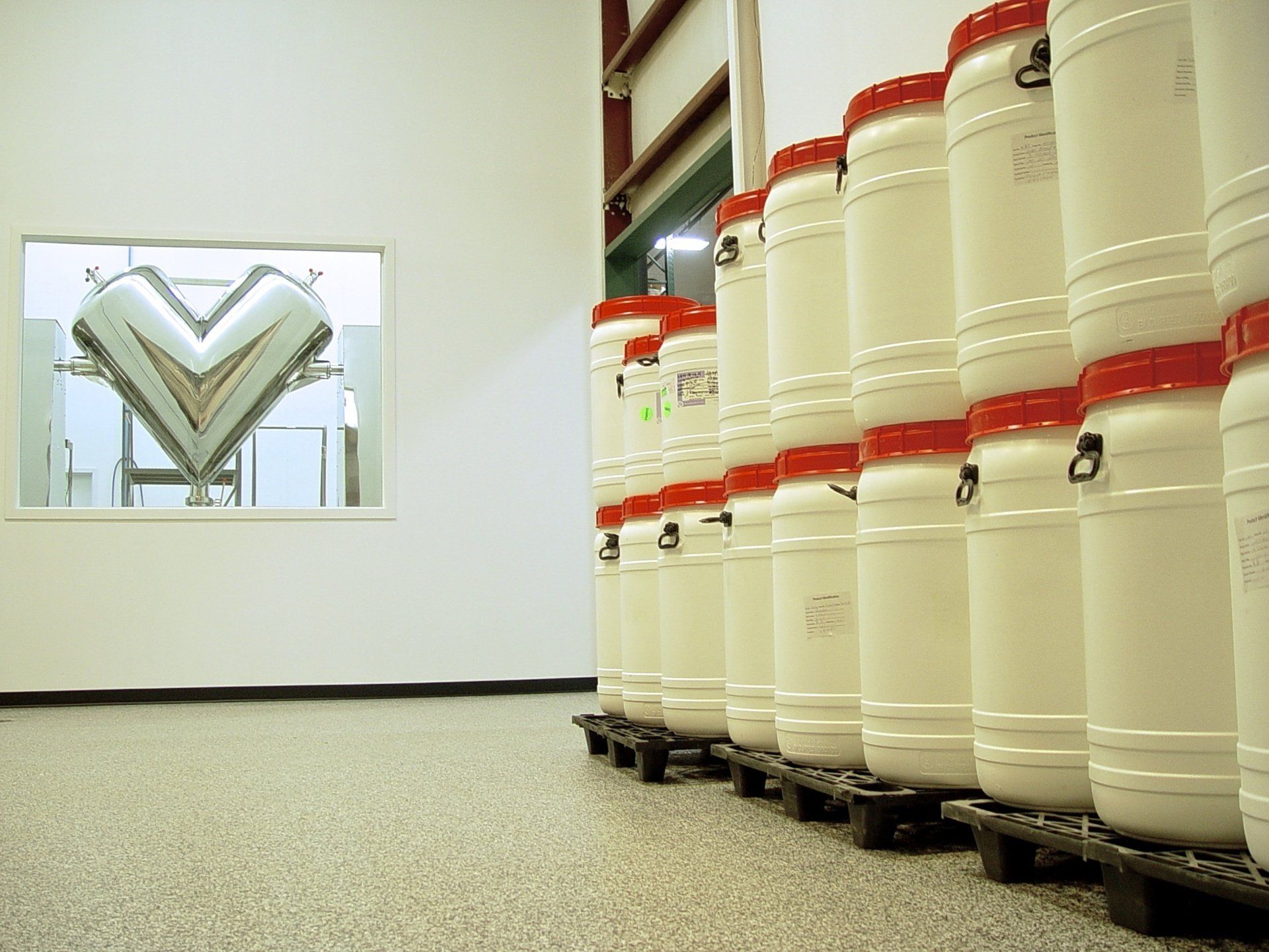 White barrels with red lids stacked on pallets in a warehouse, with a mixing machine visible through a window.