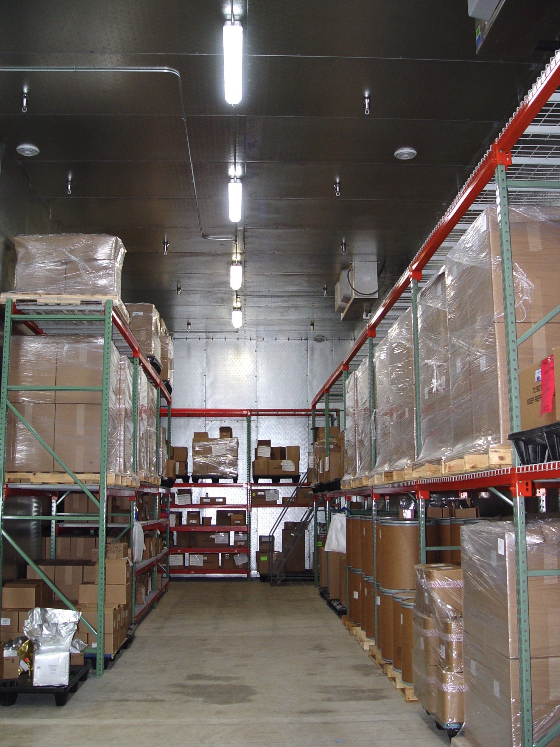 Warehouse interior with tall metal shelves holding boxed items. Fluorescent lights illuminate the aisle.