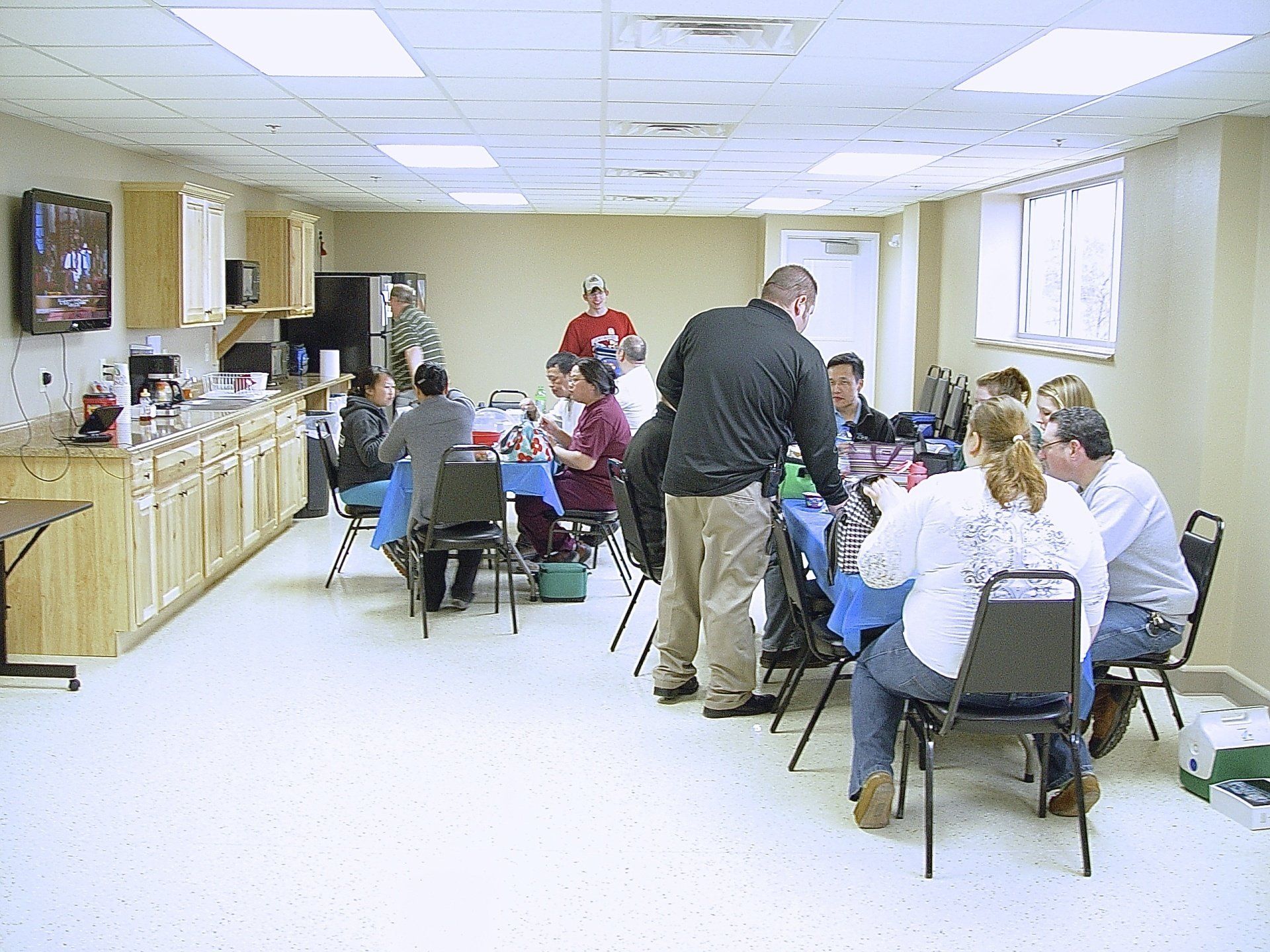 People seated at tables in a room with a kitchenette; a man serves food.