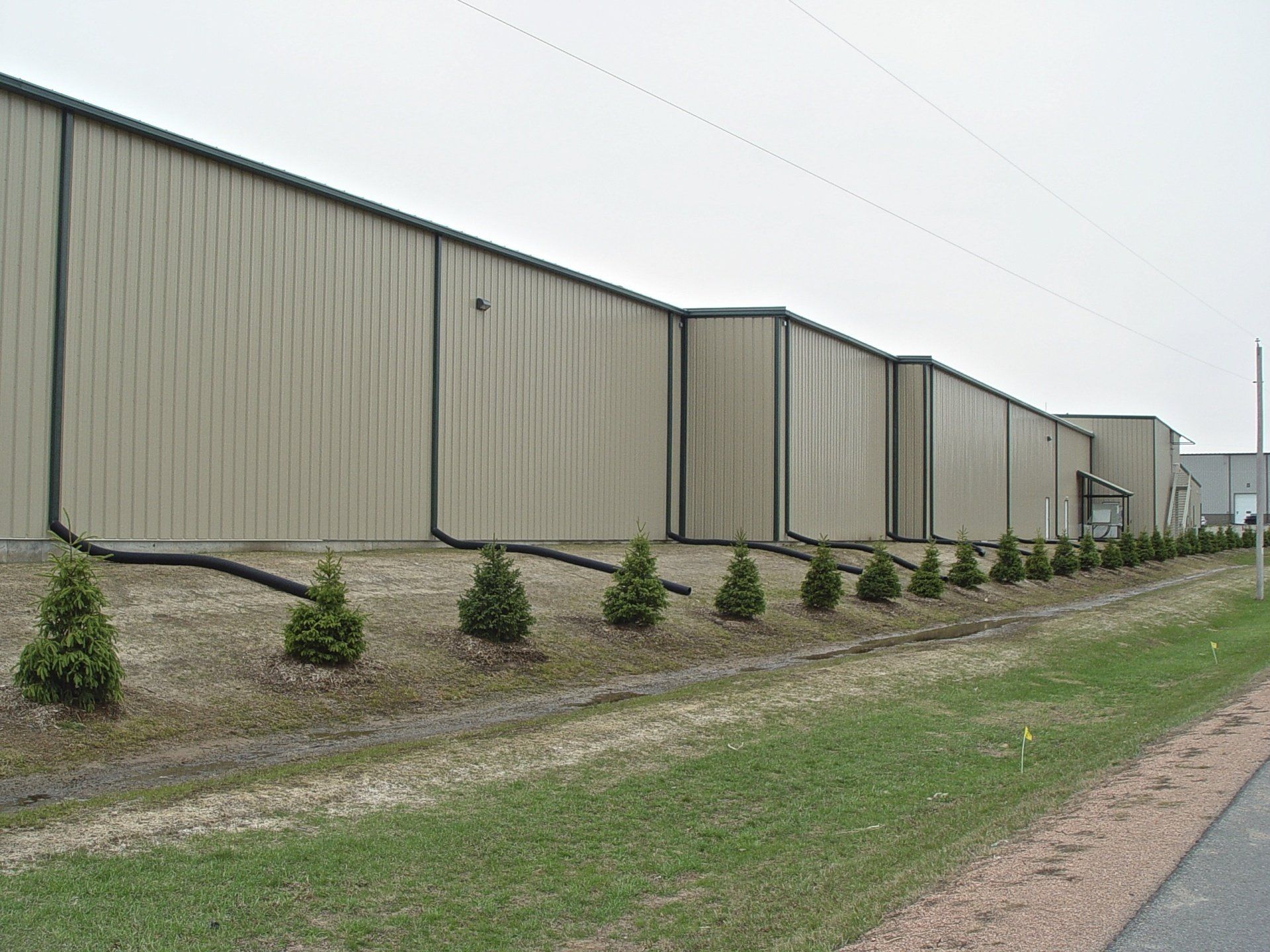Tan industrial building with black trim and small evergreen trees along the base.