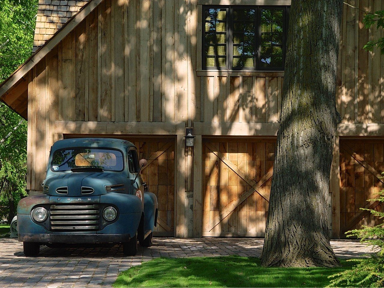 Old teal pickup truck parked in front of a rustic wooden garage with a tree in the foreground.