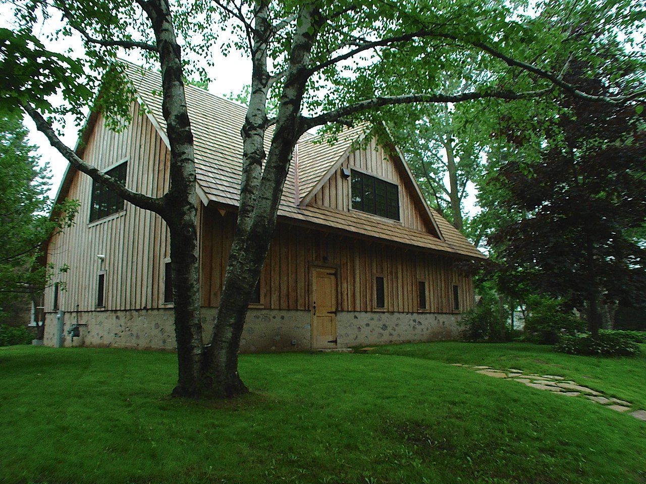 Wooden building with a gabled roof and two windows, set in a grassy yard, partially obscured by a tree.