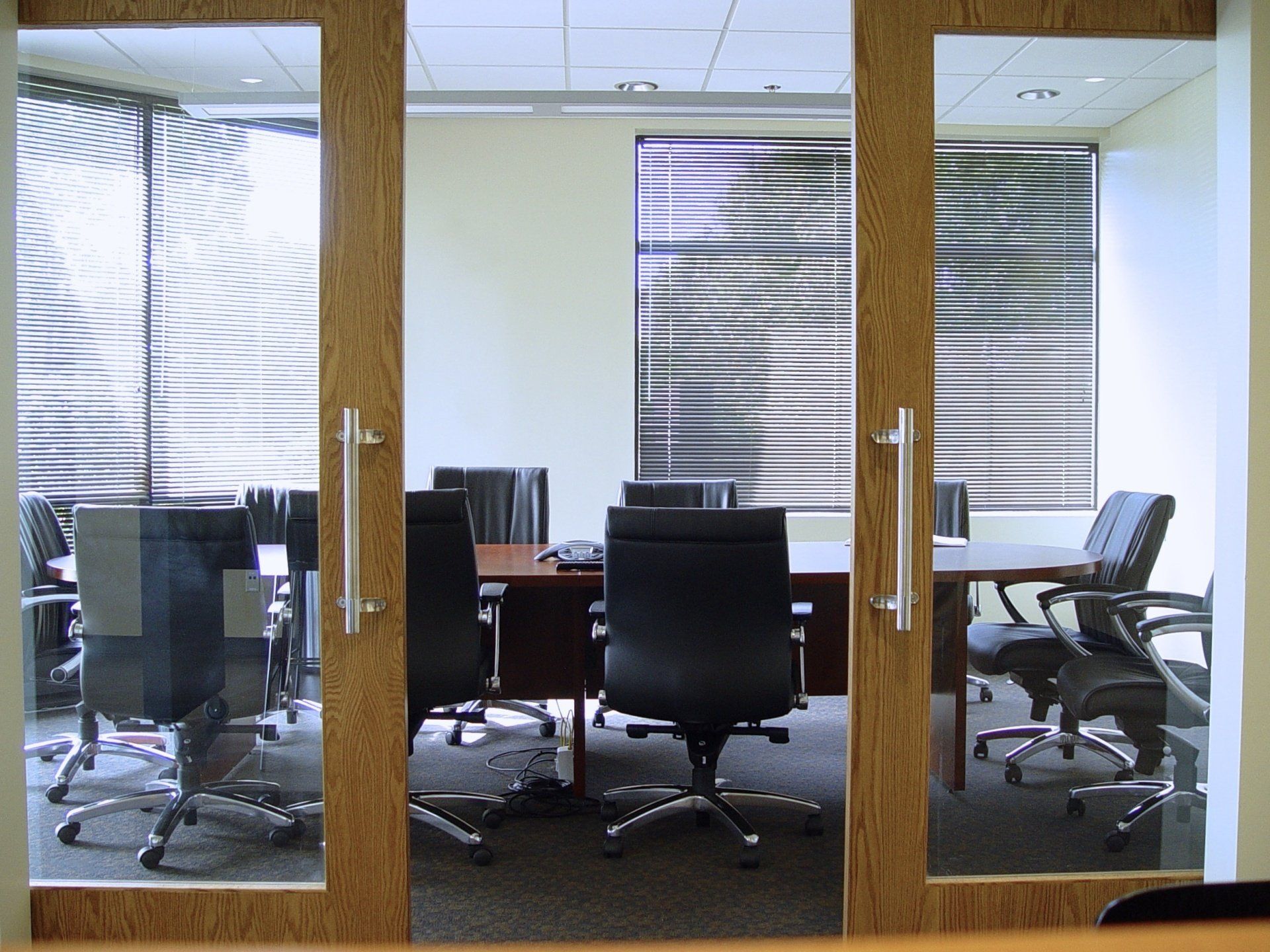 A conference room seen through double doors. Black chairs surround a brown table. Blinds cover the windows.