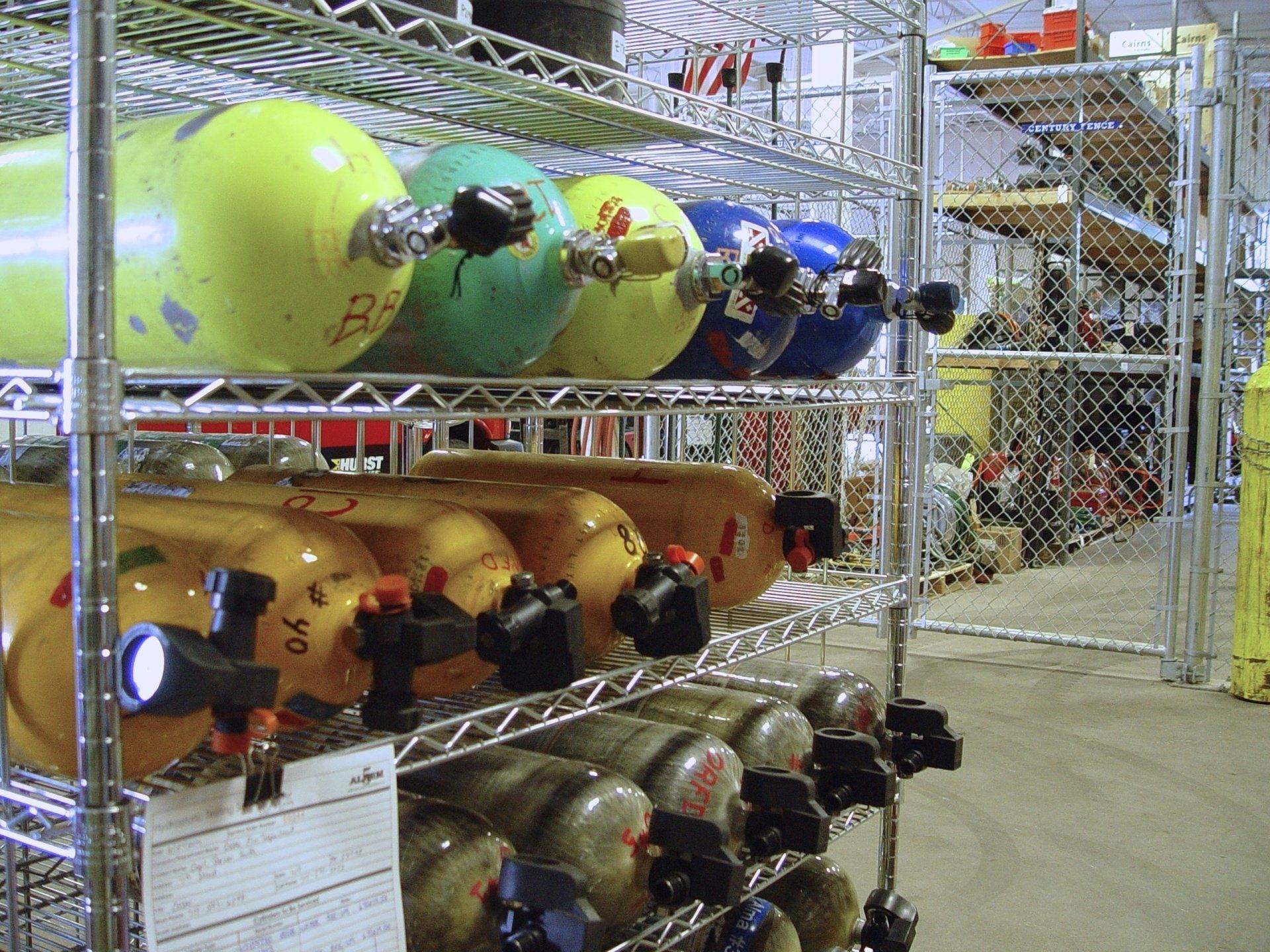 Metal shelving unit with rows of colored scuba tanks stored in a warehouse setting.