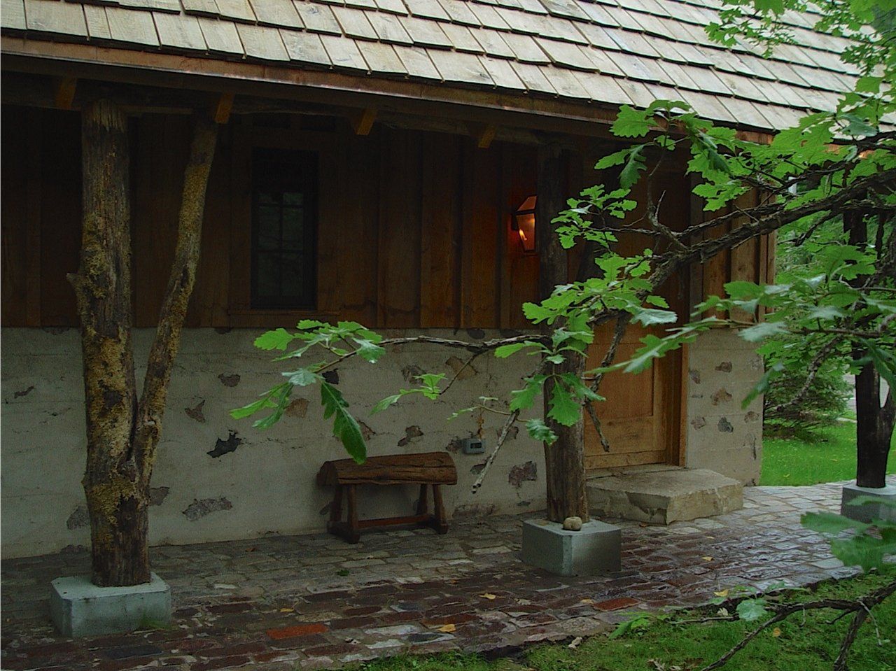 Wooden cabin with stone foundation, porch with tree-trunk pillars, bench, and green foliage.
