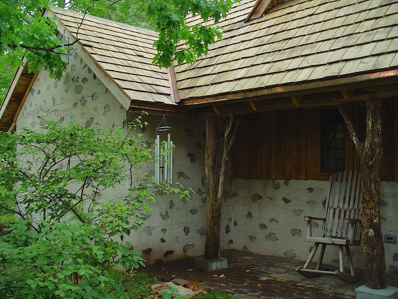 Stone-walled cabin with wood shake roof and porch; rocking chair sits outside, wind chimes hang.
