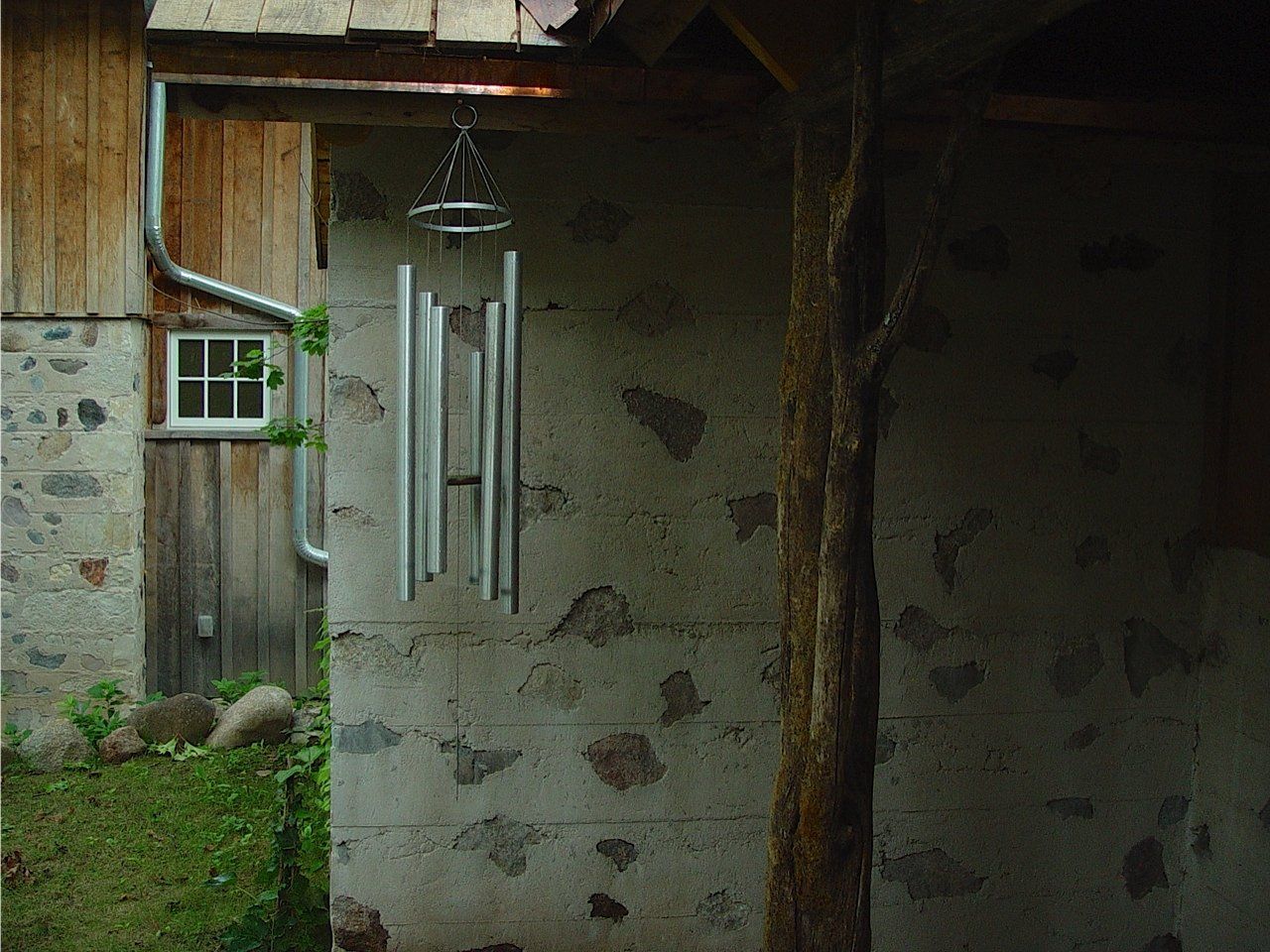 Wind chimes hang near a rustic stone and wood building with a small window and a downspout.