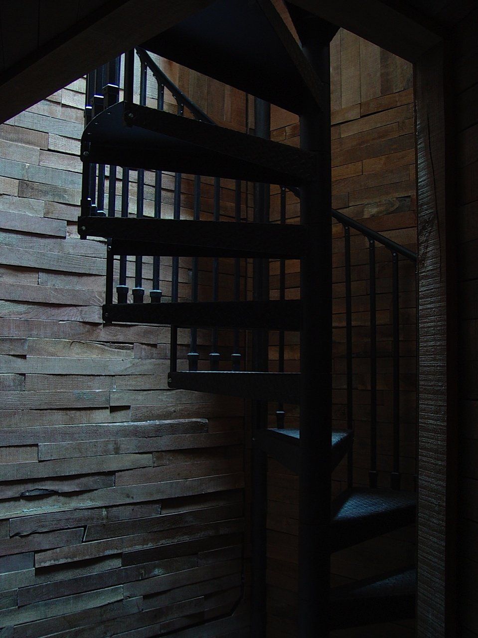 Spiral staircase in a wooden structure. Dark metal railing, steps. Wooden wall panels. Dim lighting.
