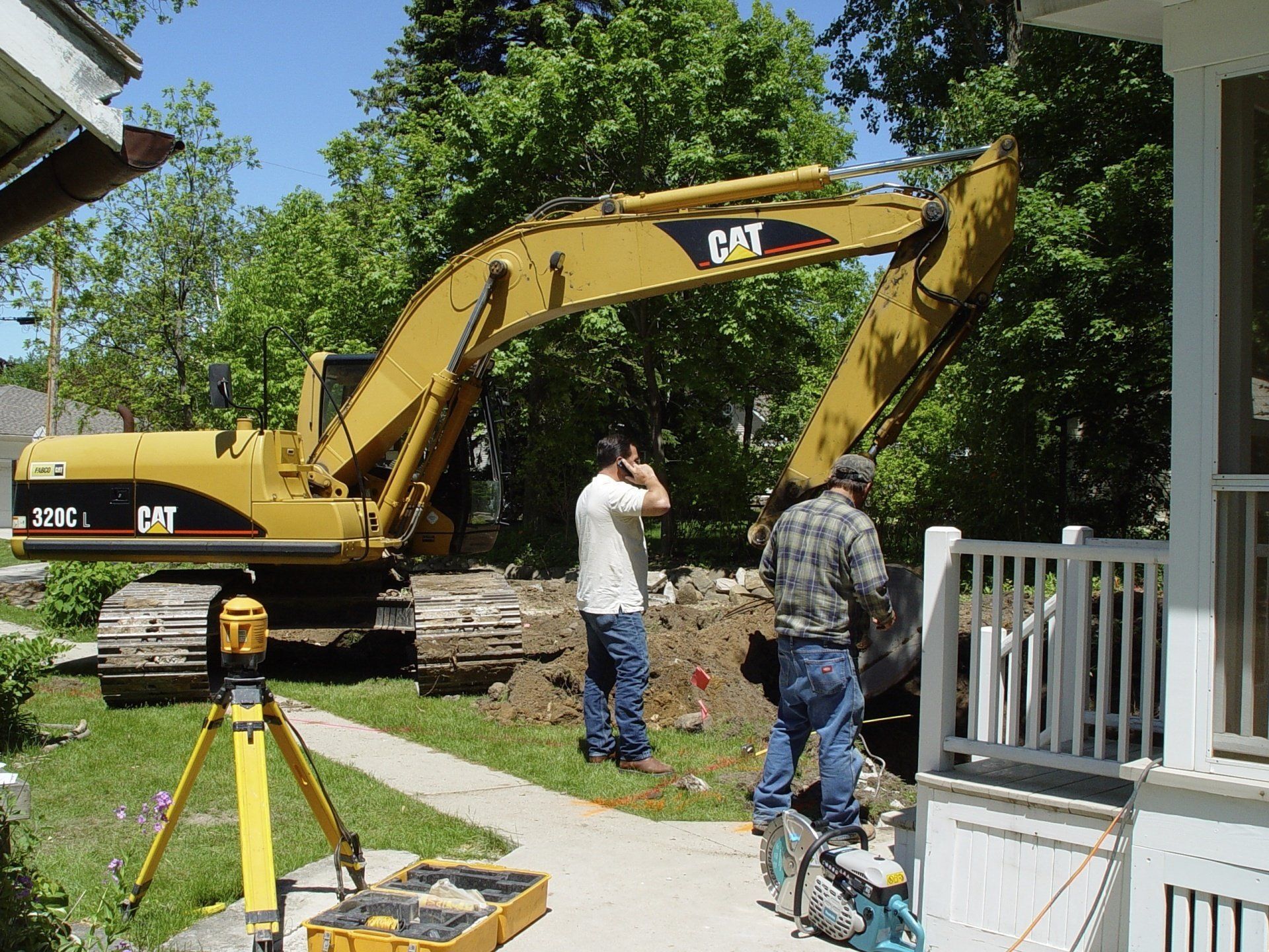 Yellow CAT excavator digging near a house; two men watch. Survey equipment in foreground. Sunny outdoor setting.
