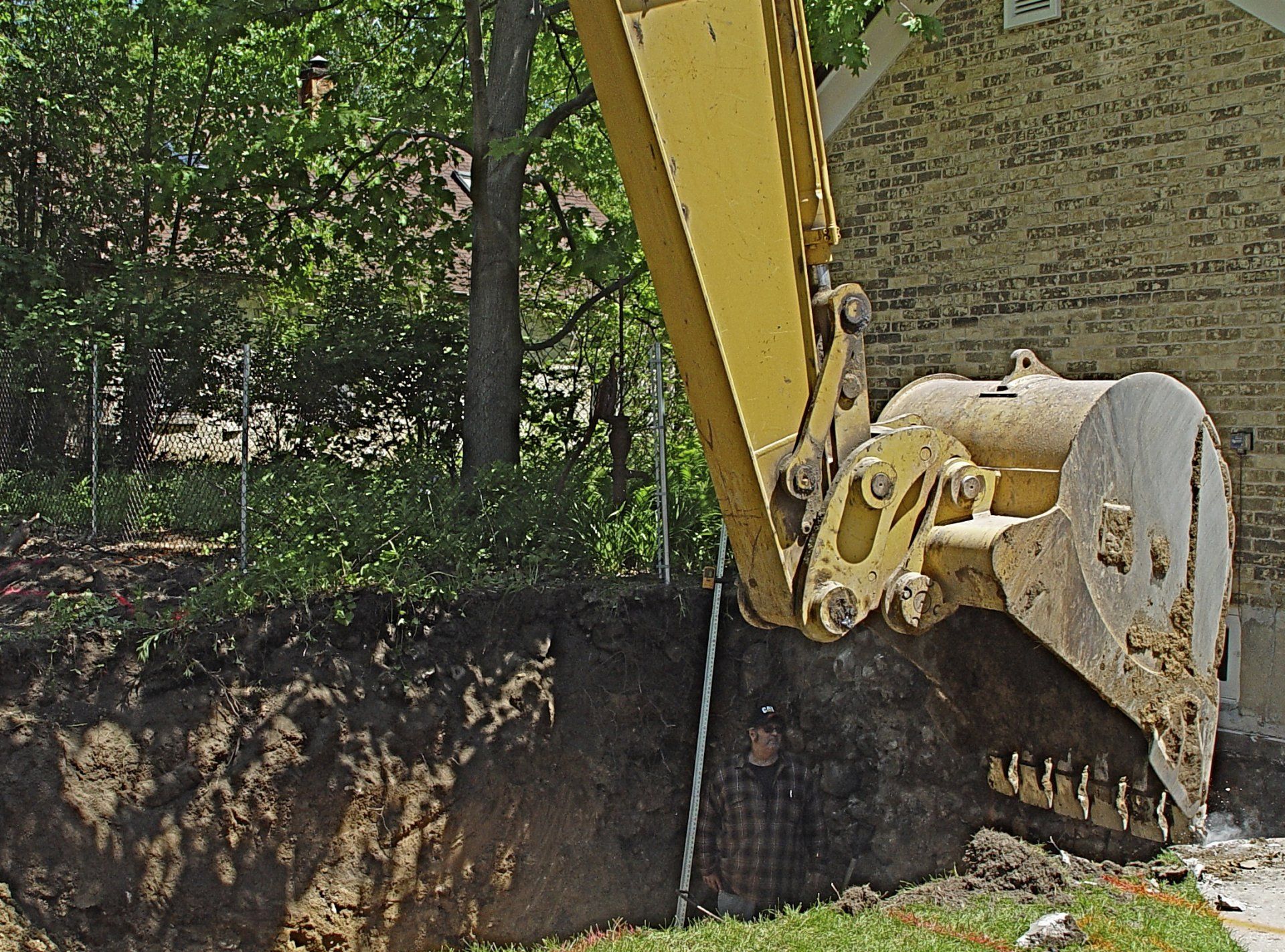 Yellow excavator digging into a dirt bank near a house. Green trees and grass are in the background.