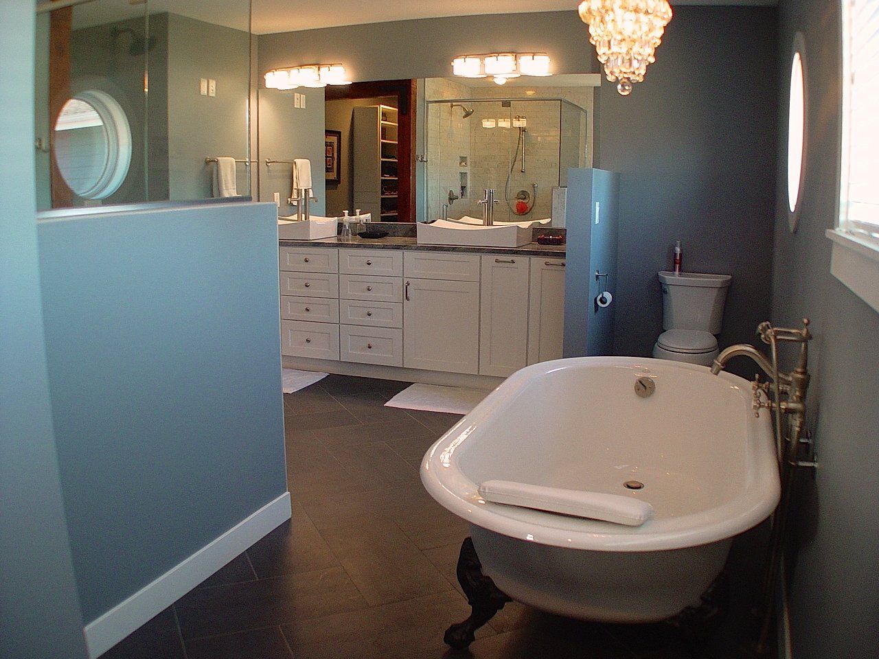 Elegant bathroom with clawfoot tub, white cabinetry, and chandelier. Dark floor, blue walls, and large mirror.