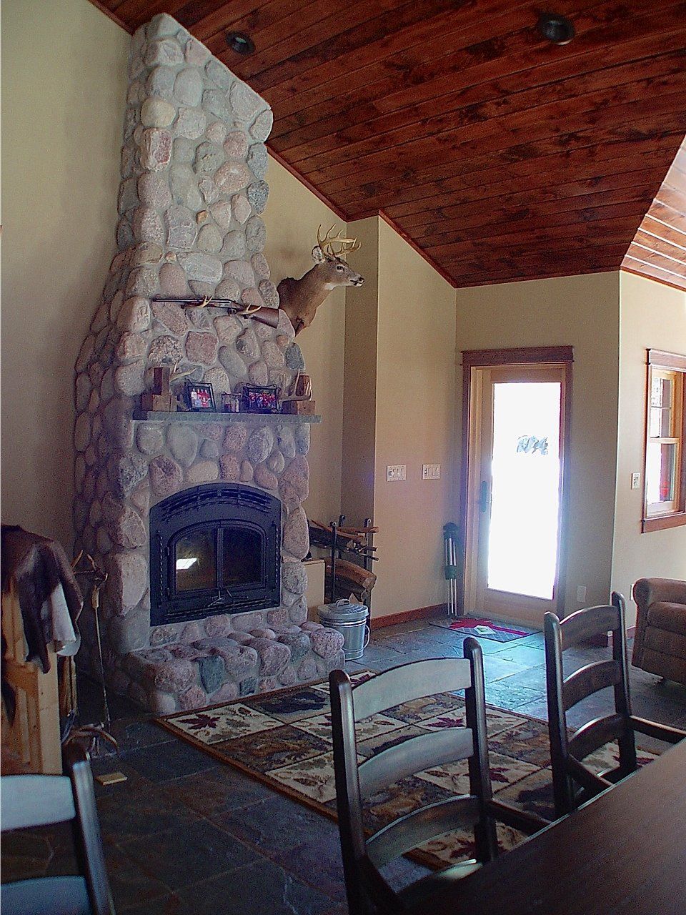 Stone fireplace in a cabin interior, mounted deer head, wooden ceiling and door, dining chairs, area rug.
