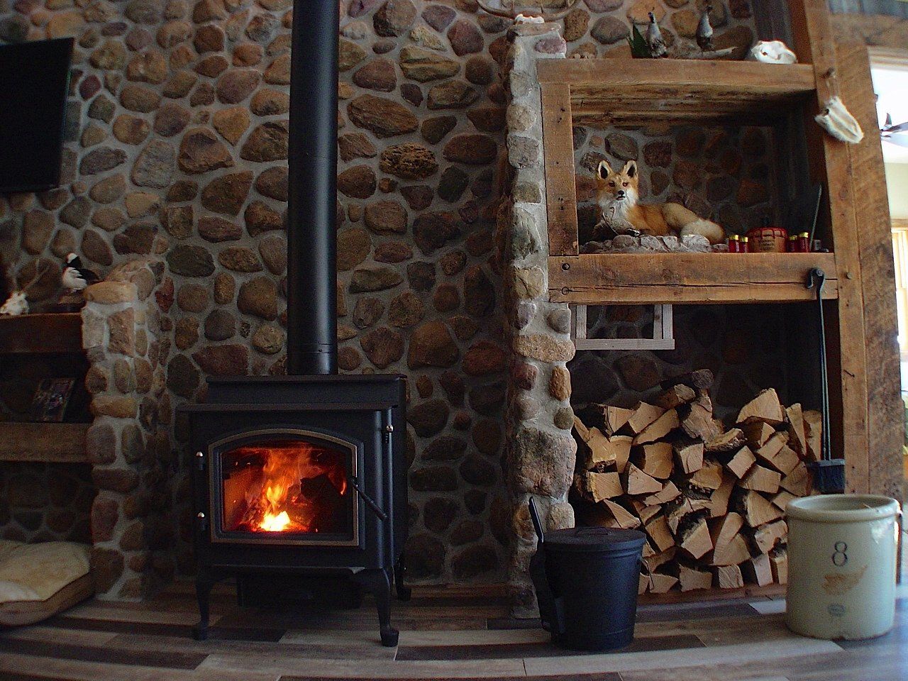 Fireplace with wood-burning stove against a stone wall. Fox figurine on a shelf above firewood.
