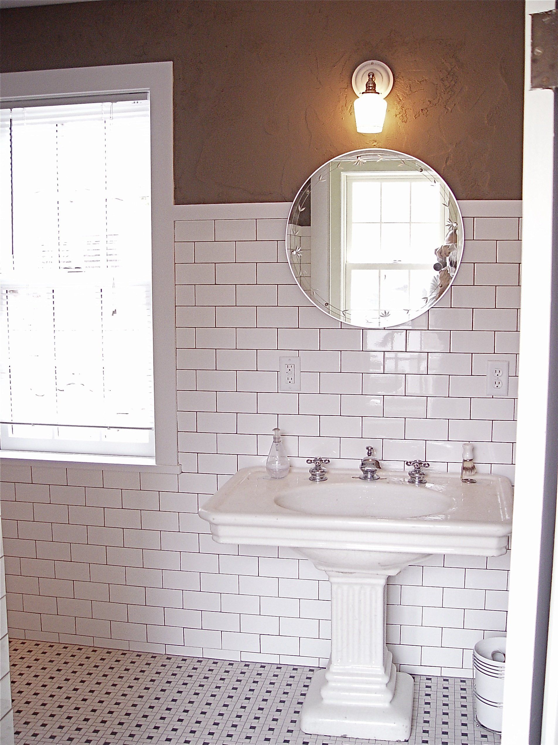 Bathroom with white tiled walls, pedestal sink, round mirror, window, and patterned floor.