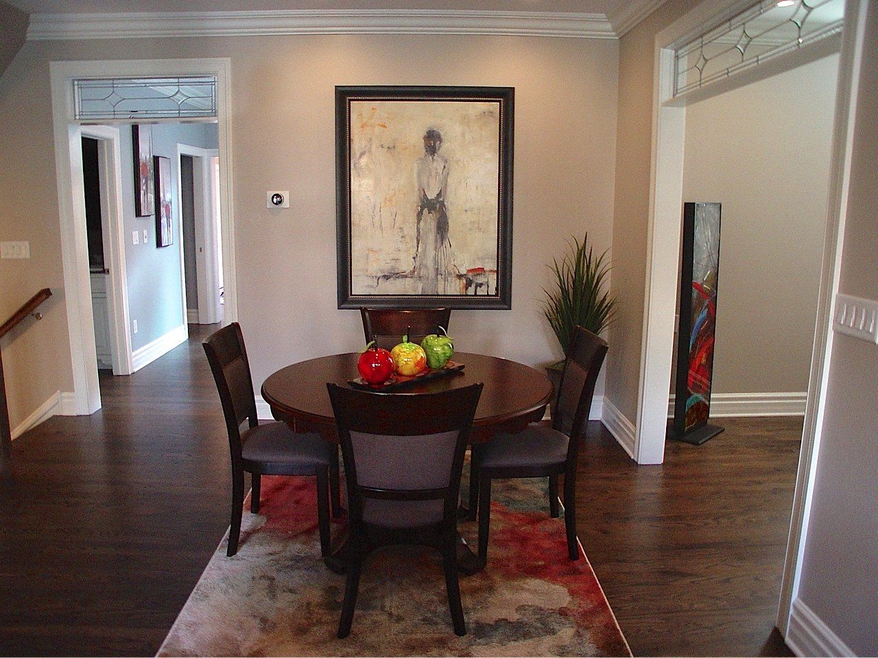 Dining room with round table, chairs, and painting. Rug and dark wood floor. Two doorways visible.