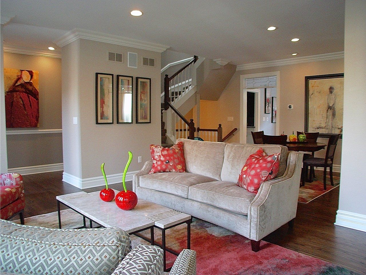 Living room with sofa, artwork, and staircase. Beige walls, wood floors, and red accents.