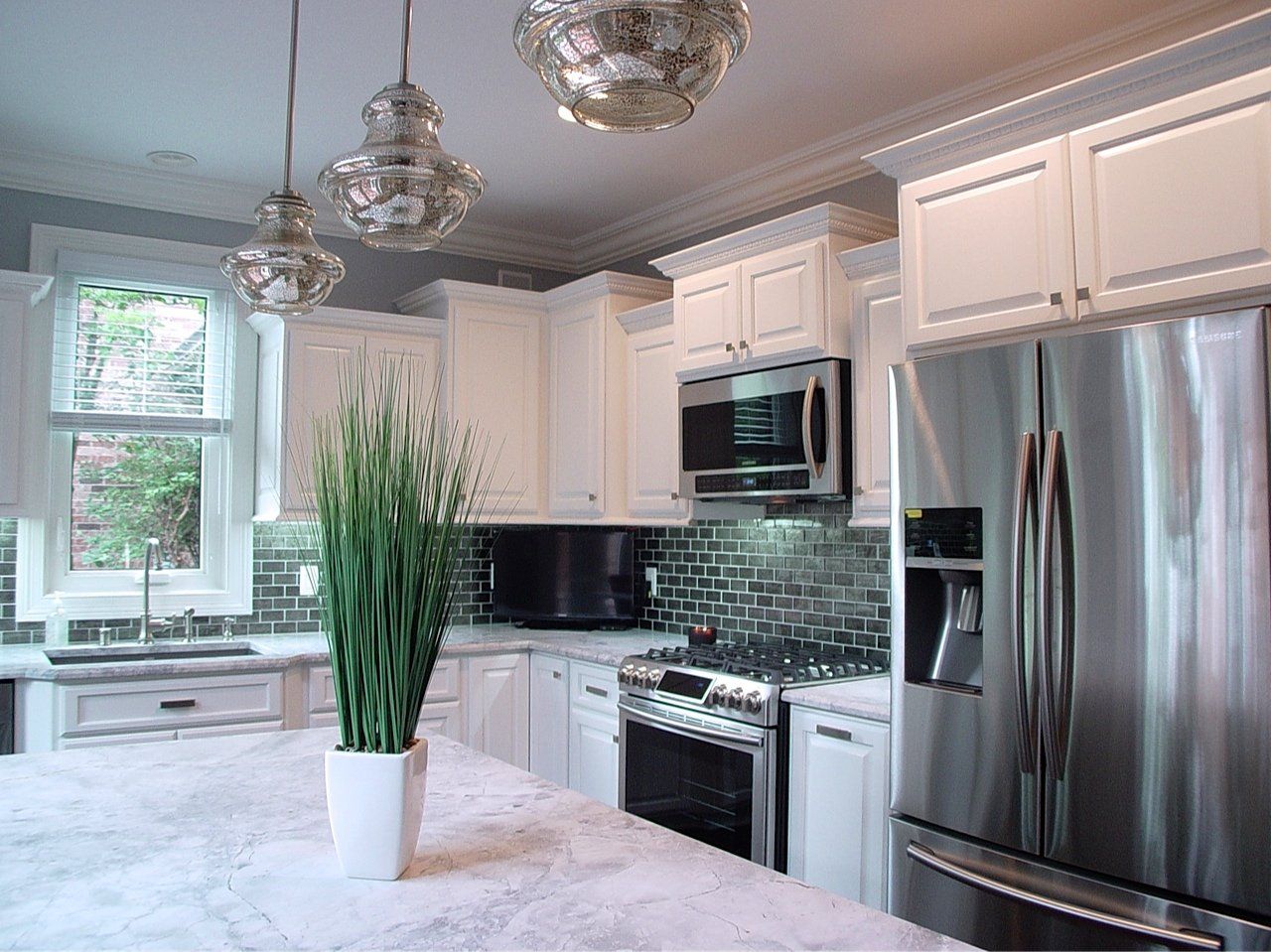 Modern white kitchen with stainless steel appliances, decorative lights, and a plant on the island.