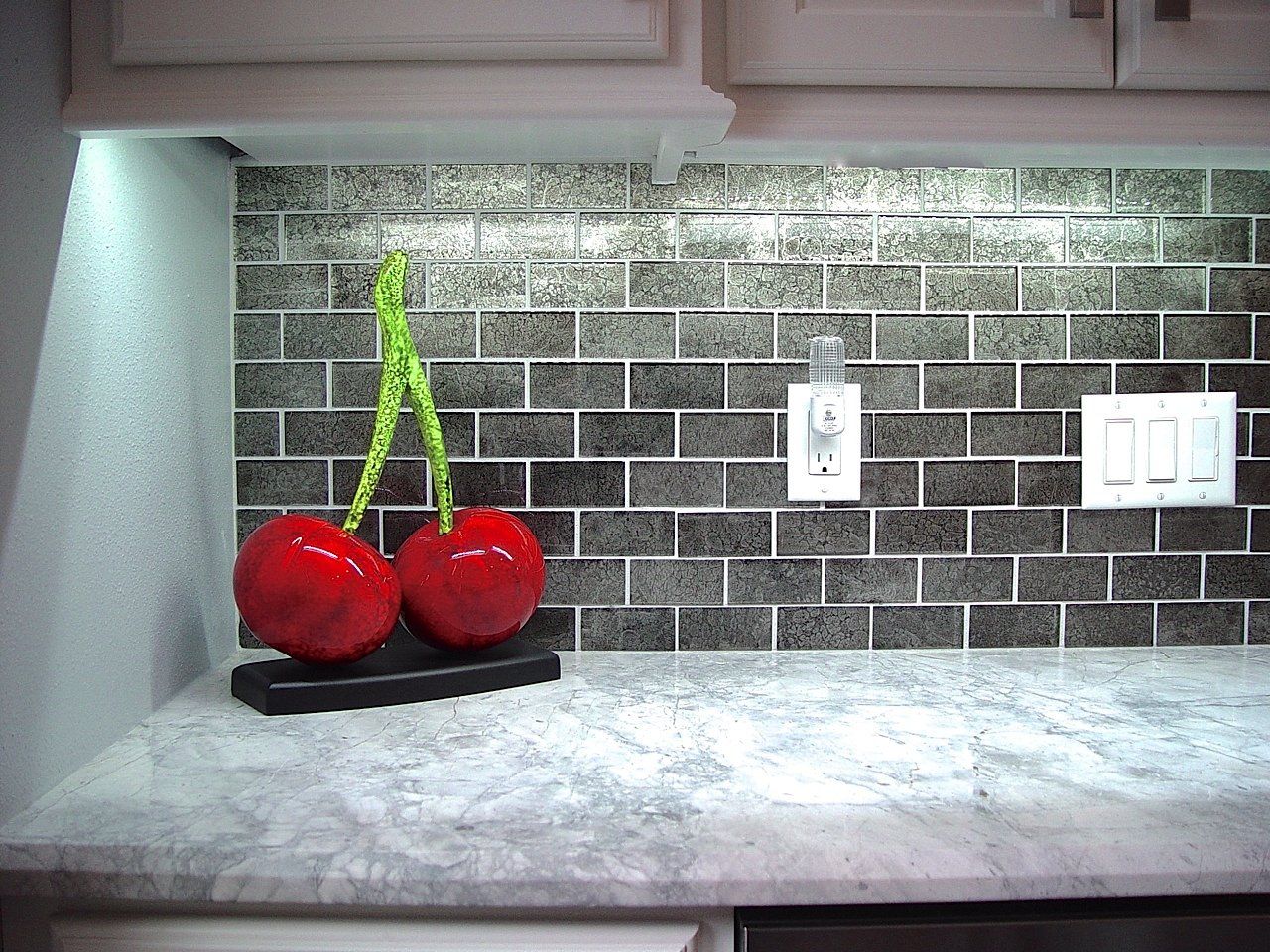 Cherries on countertop. Gray tile backsplash, white cabinets, outlet, and light switches.