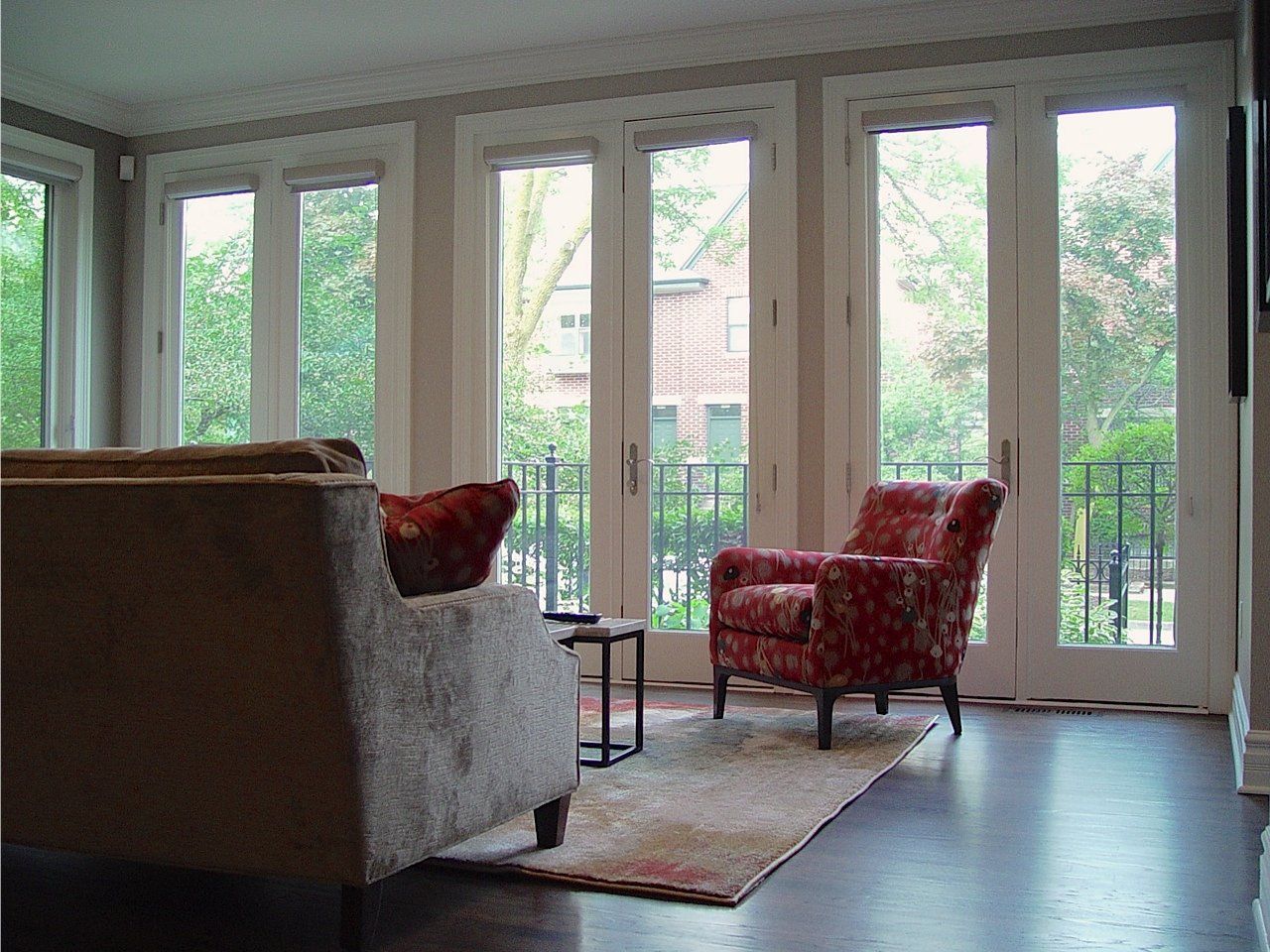 Living room with large windows overlooking a yard, brown couch, floral armchair, small table, rug.