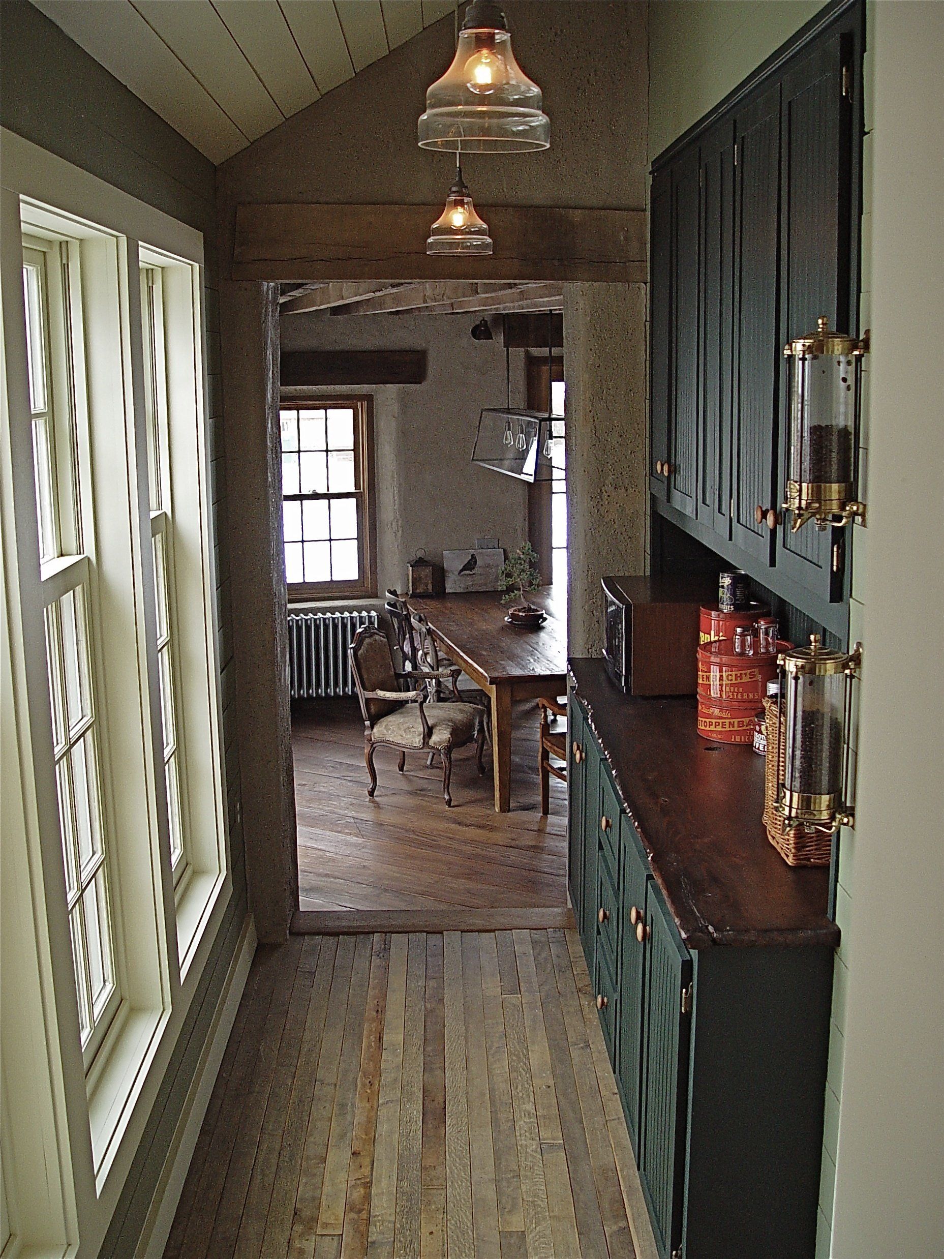 Narrow hallway leading to a dining area. Dark green cabinets, wooden floors, and glass pendant lights.