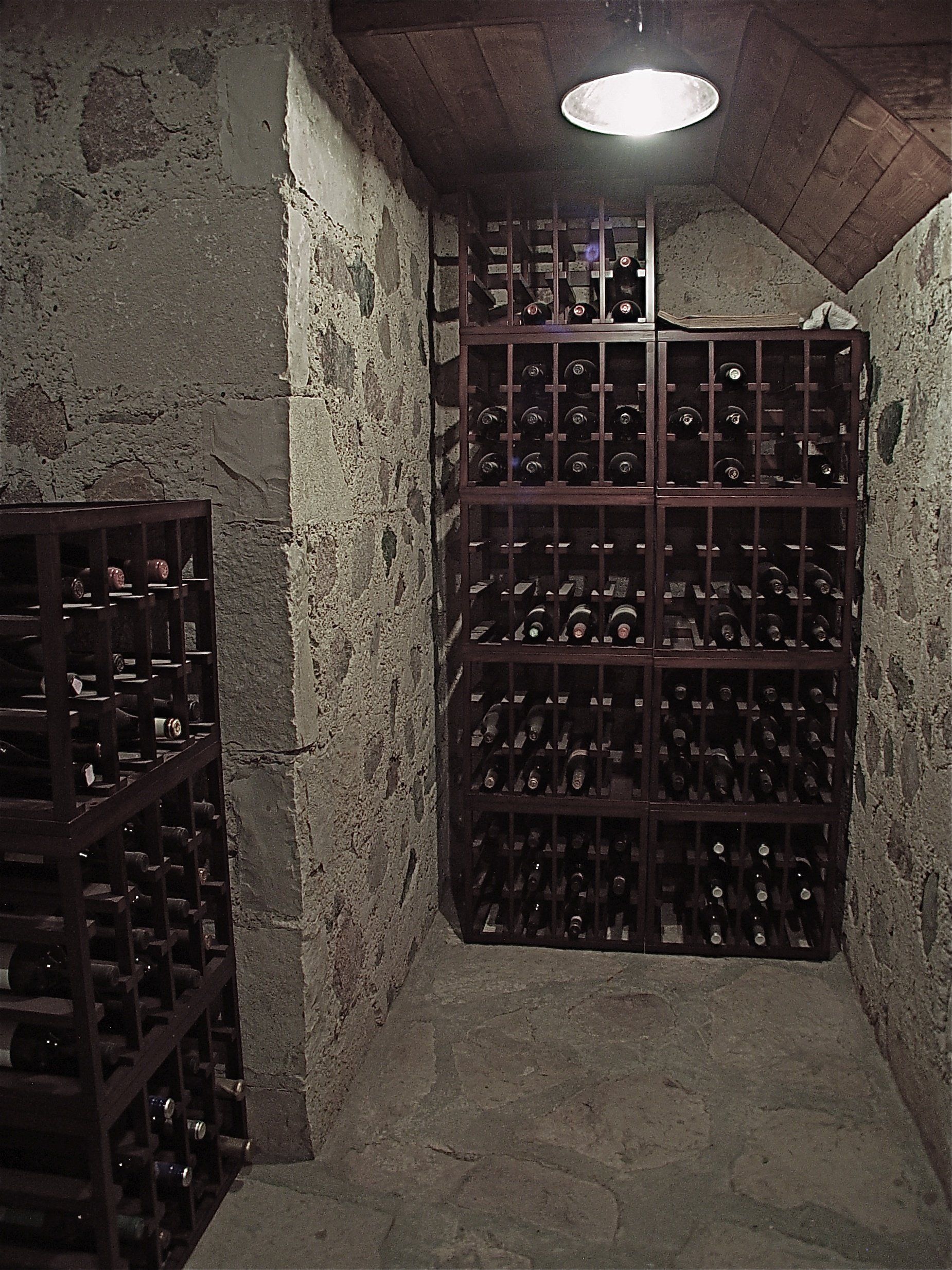 Wine cellar with stacked wooden racks filled with bottles, set in a stone-walled room under a ceiling light.