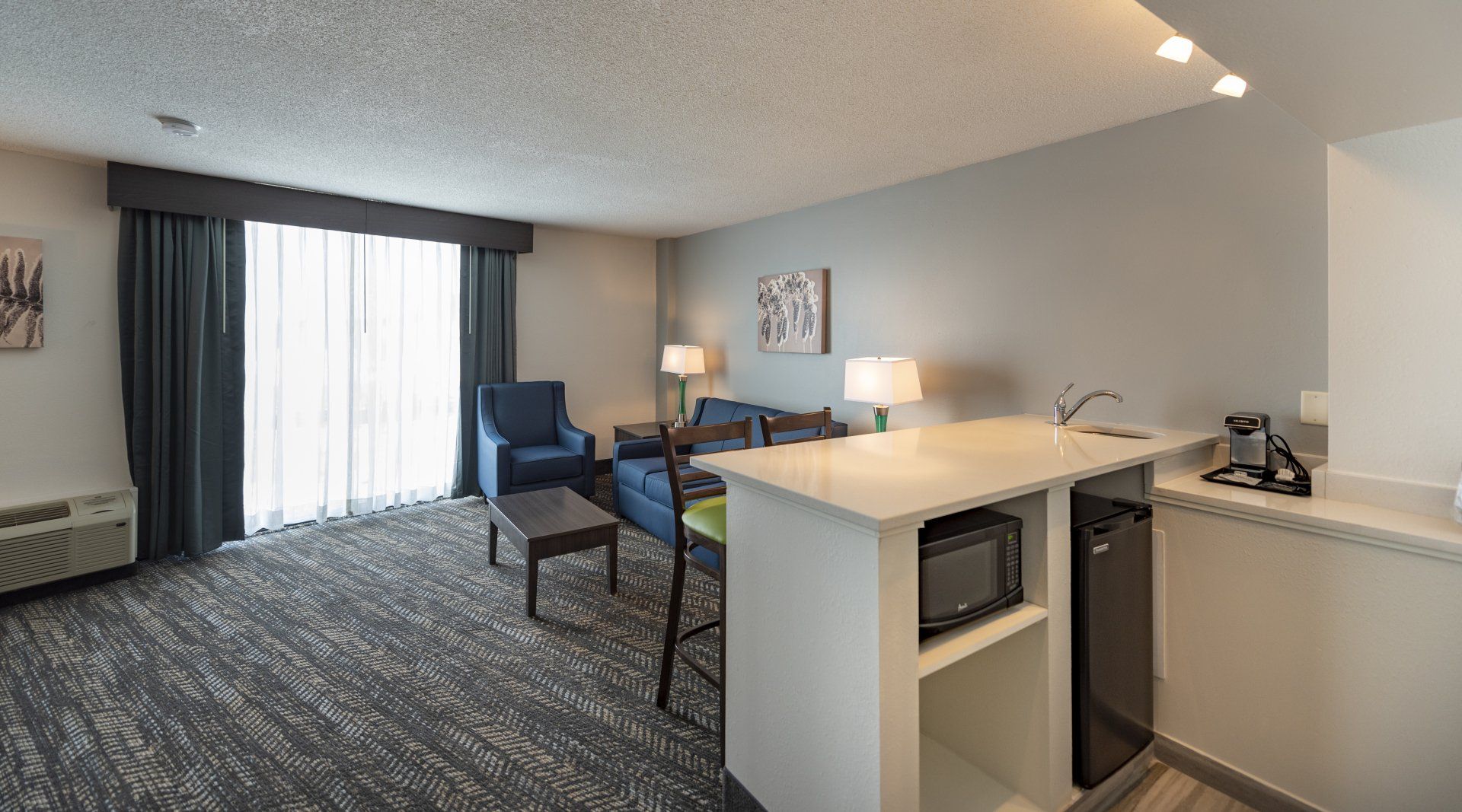 Hotel suite living area: gray walls, patterned carpet, sofa, kitchenette with microwave and mini-fridge, large window with curtains.