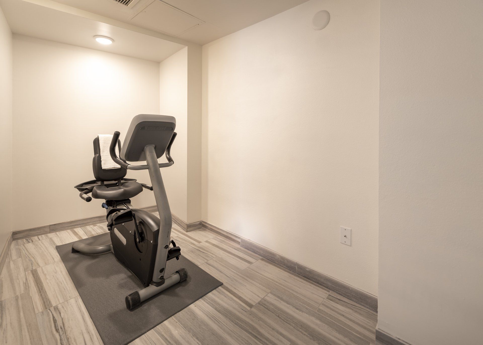 Recumbent exercise bike on a mat in a small, white-walled room with wood-look flooring.