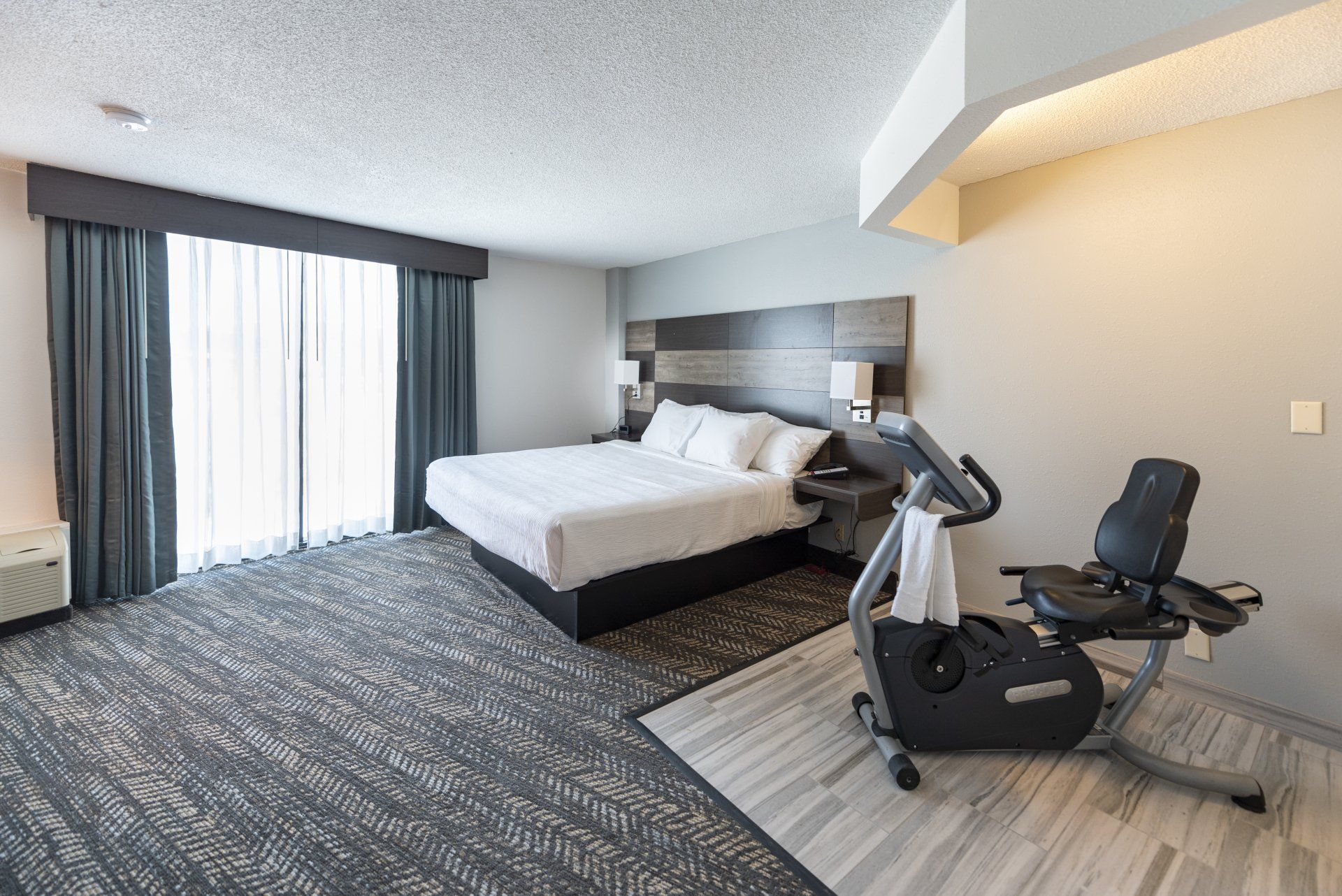 Hotel room with bed, exercise bike, and window with dark curtains. Gray carpet.